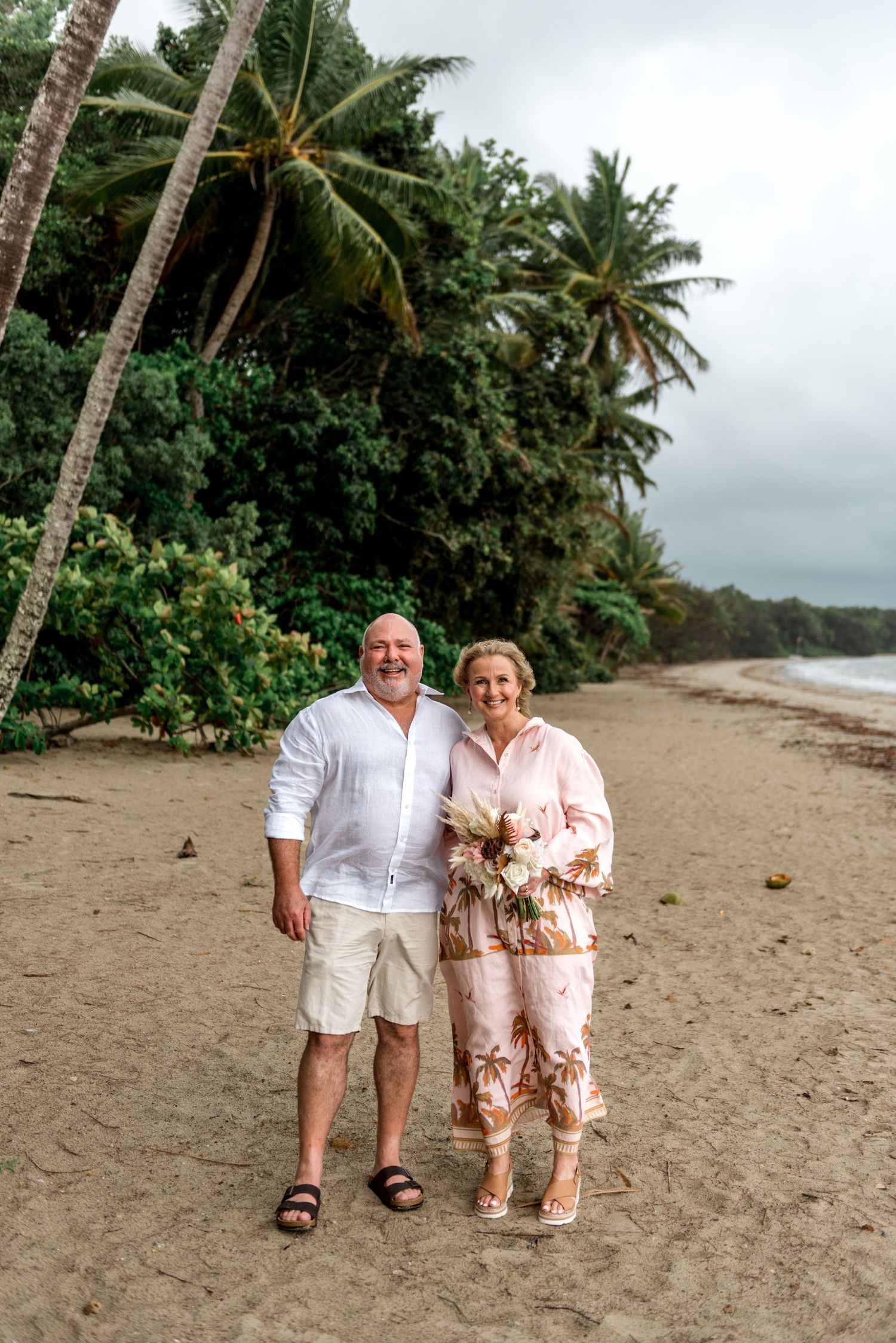 Smiling couple posing on a tropical beach with palm trees and lush greenery behind them.