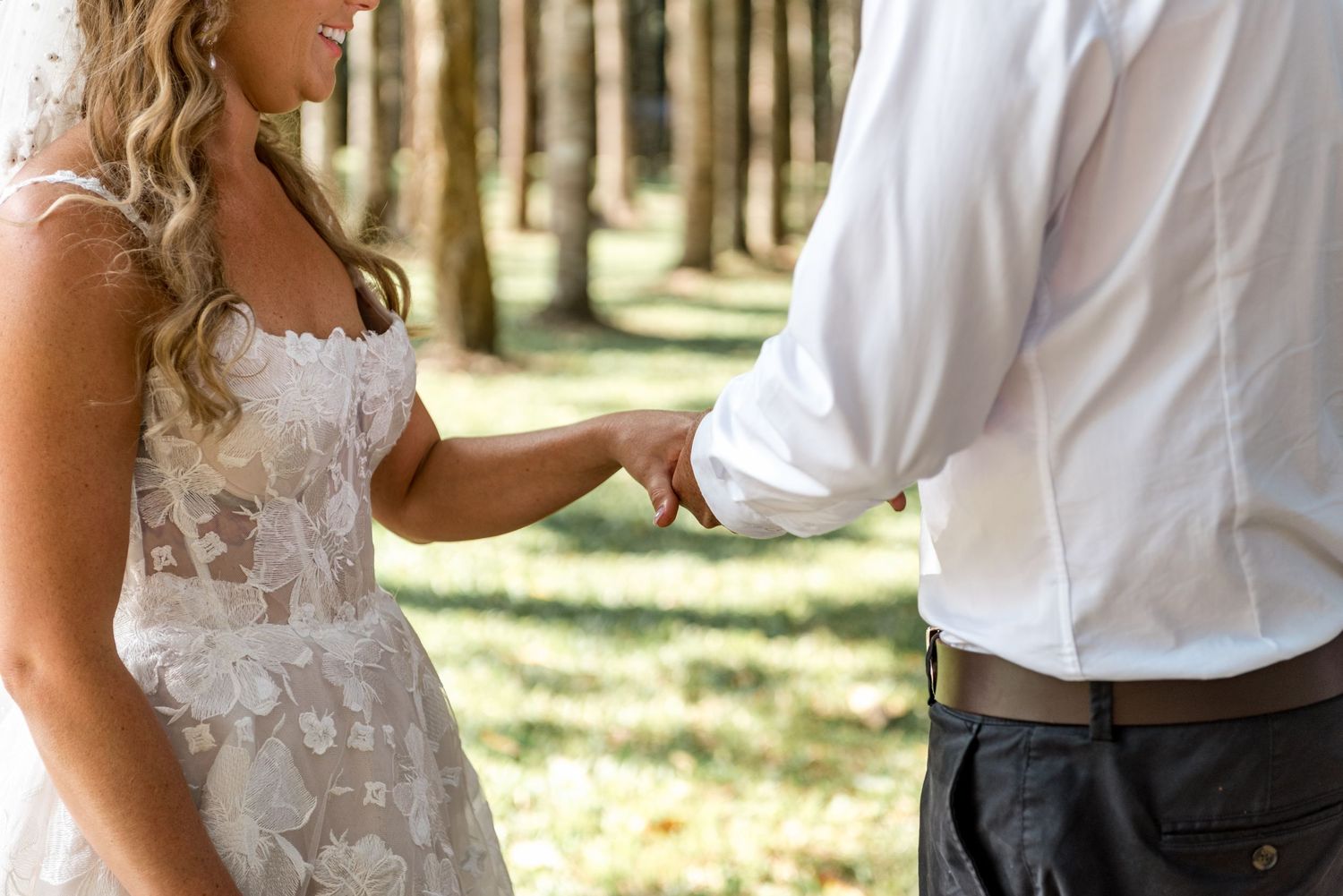 Close-up of bride in lace gown holding grooms hand during outdoor woodland wedding ceremony.