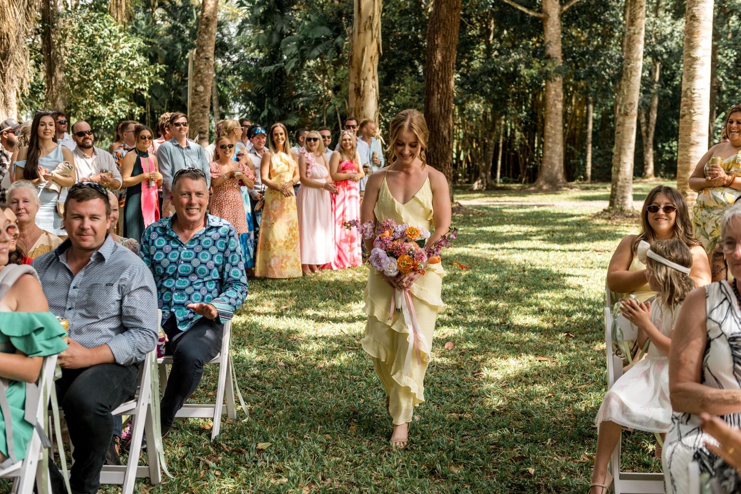 Bridesmaid walks down the aisle carrying colorful bouquet past smiling seated wedding guests.
