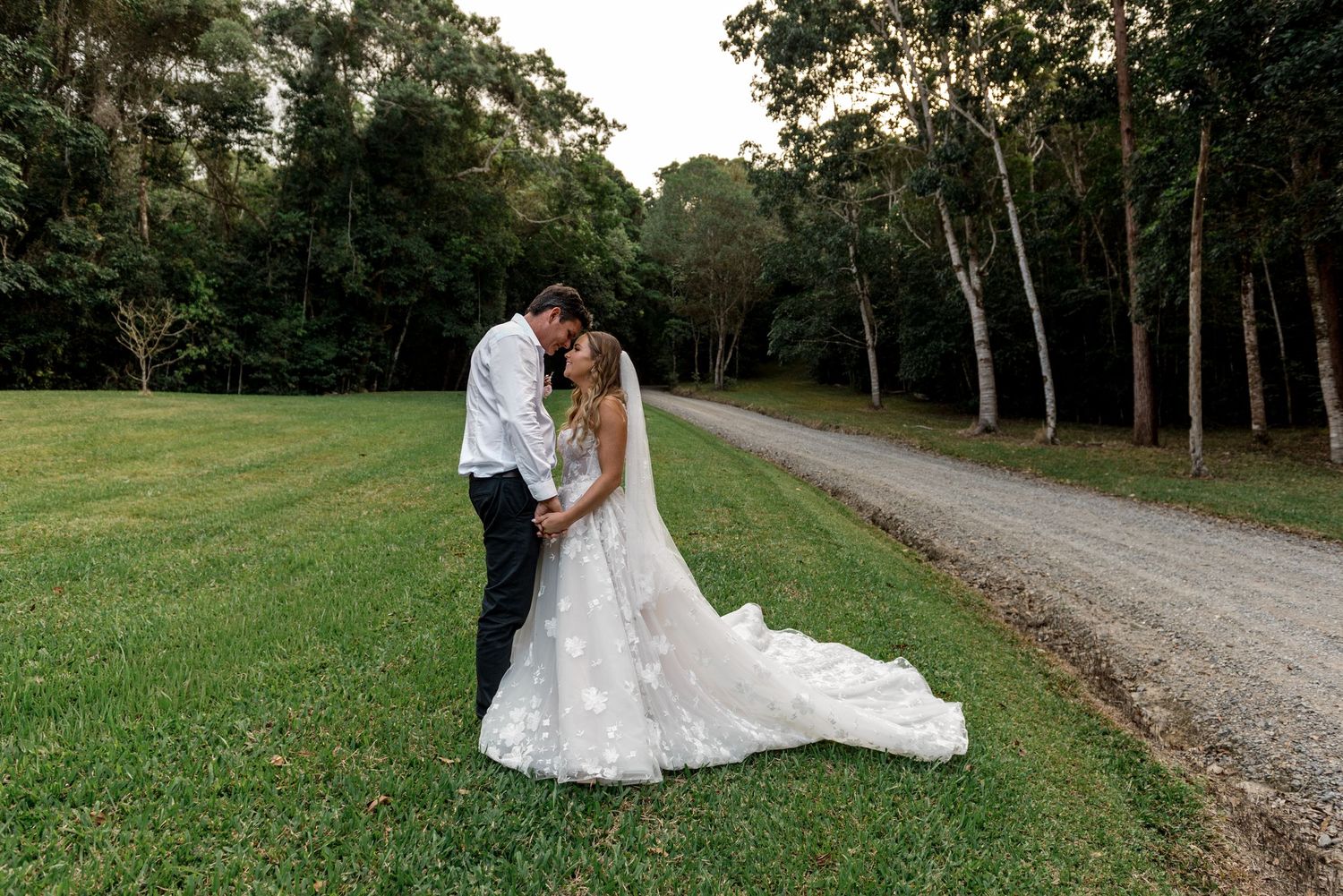 Bride and groom share a romantic kiss beside a gravel path surrounded by lush green forest at sunset.