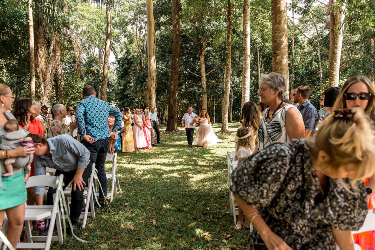 Outdoor wedding ceremony in a forest with guests seated on white chairs along a grass aisle.