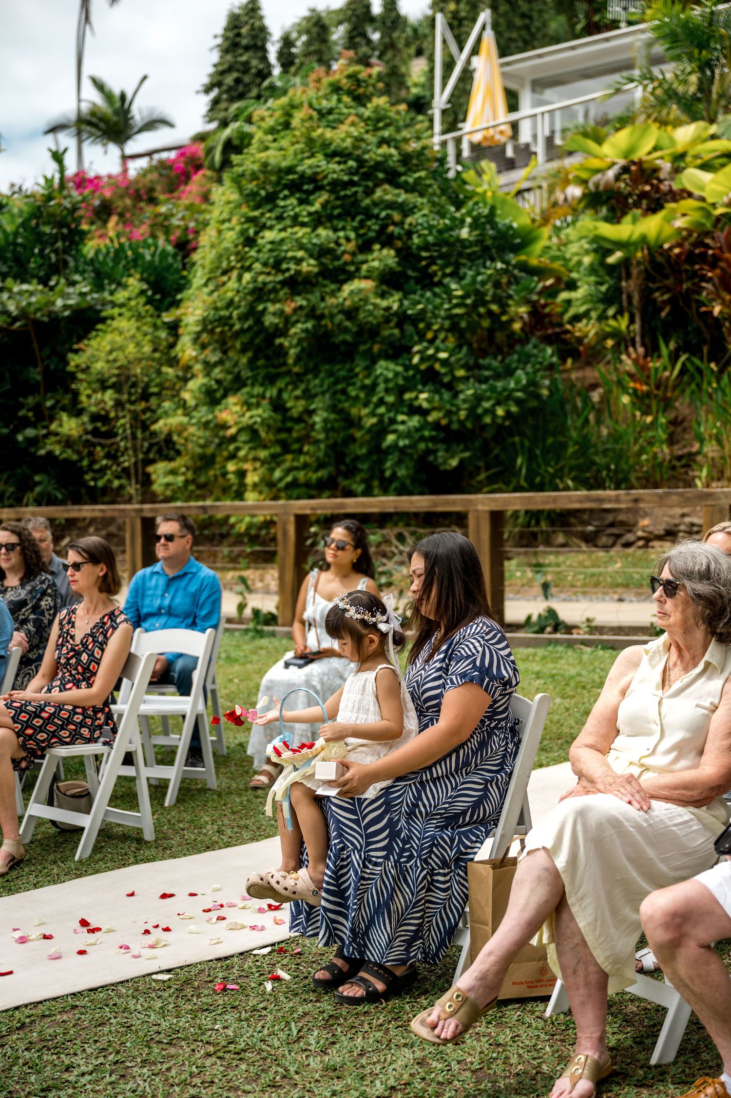 Wedding guests seated on white chairs watch a ceremony on a tropical garden lawn with rose petal aisle at Little Cove, Port Douglas.