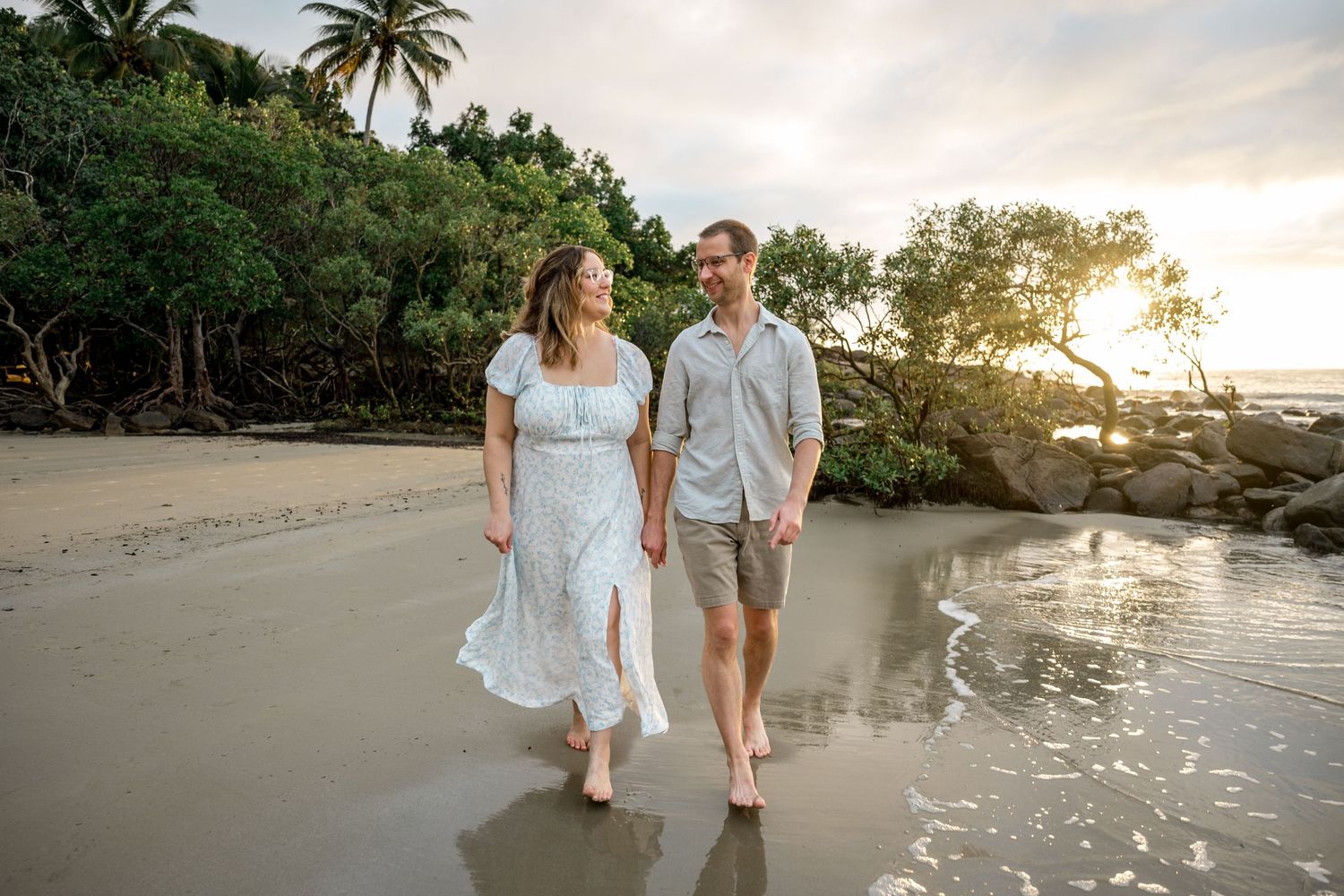 Couple walks barefoot along wet sandy beach at golden sunset with tropical mangrove trees behind at Four Mile Beach, Port Douglas.