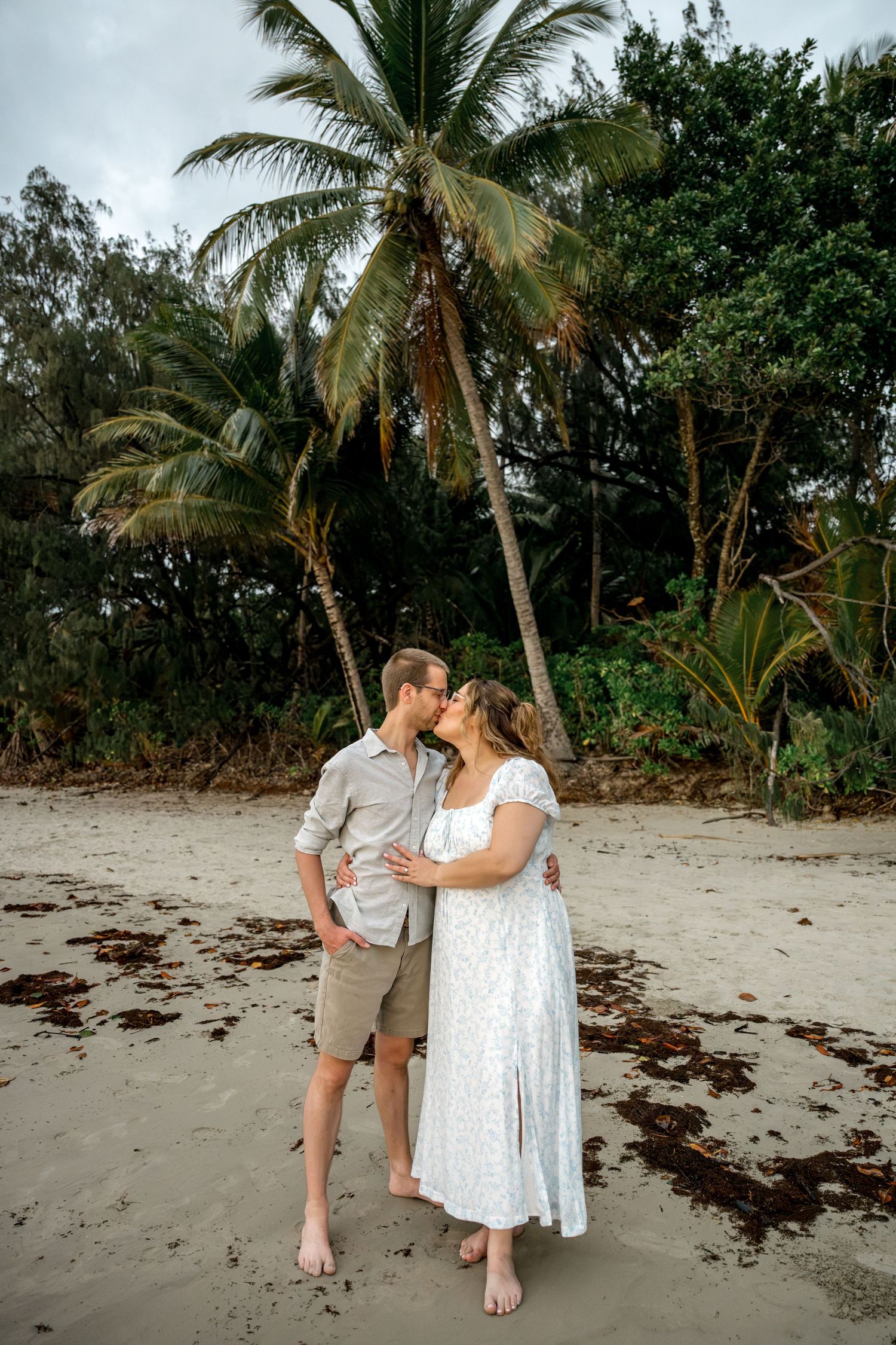 Couple sharing a tender kiss while standing barefoot on tropical beach with palm trees behind them at Four Mile Beach, Port Douglas.