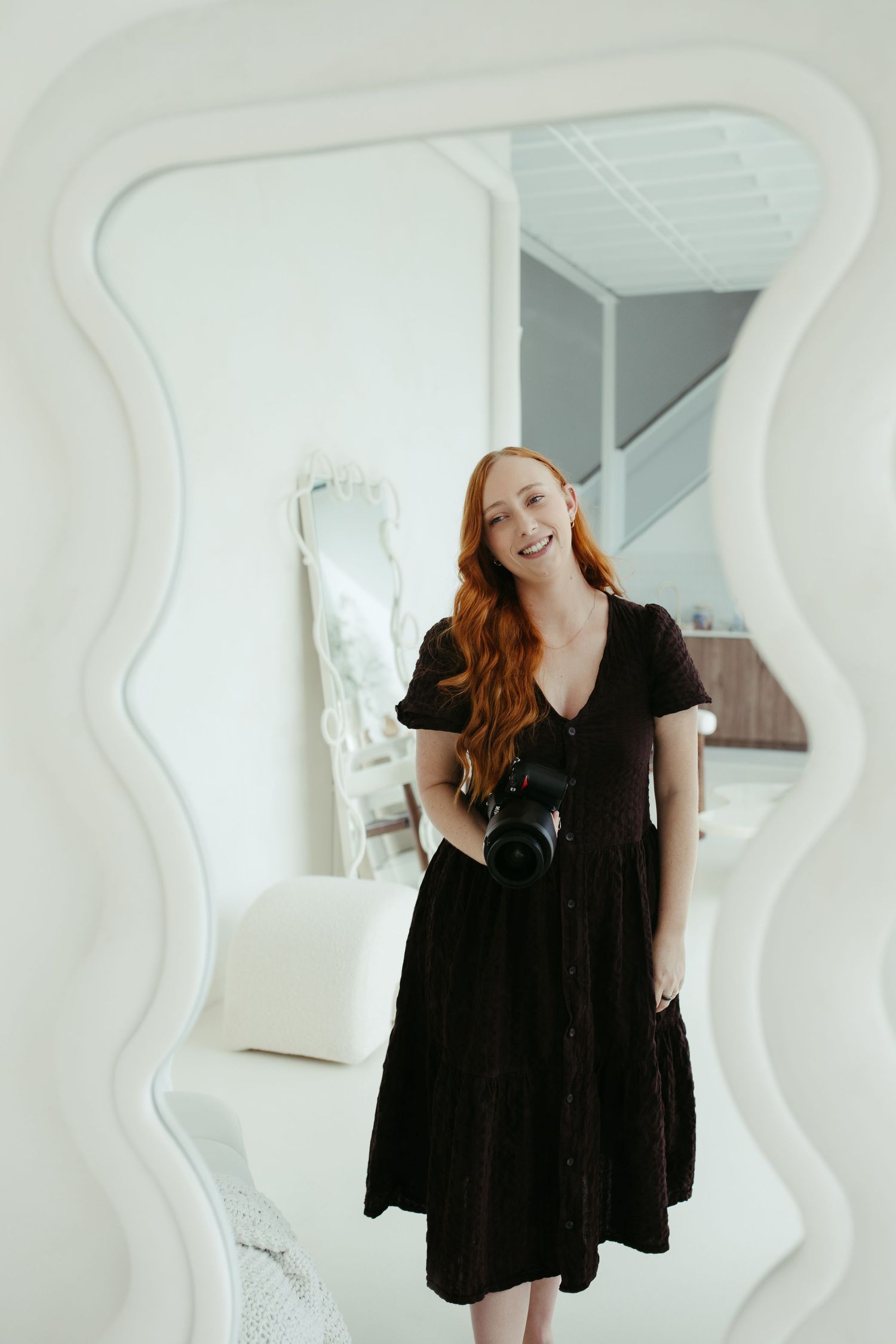 Smiling redhead woman in black dress holding camera, reflected in decorative mirror.