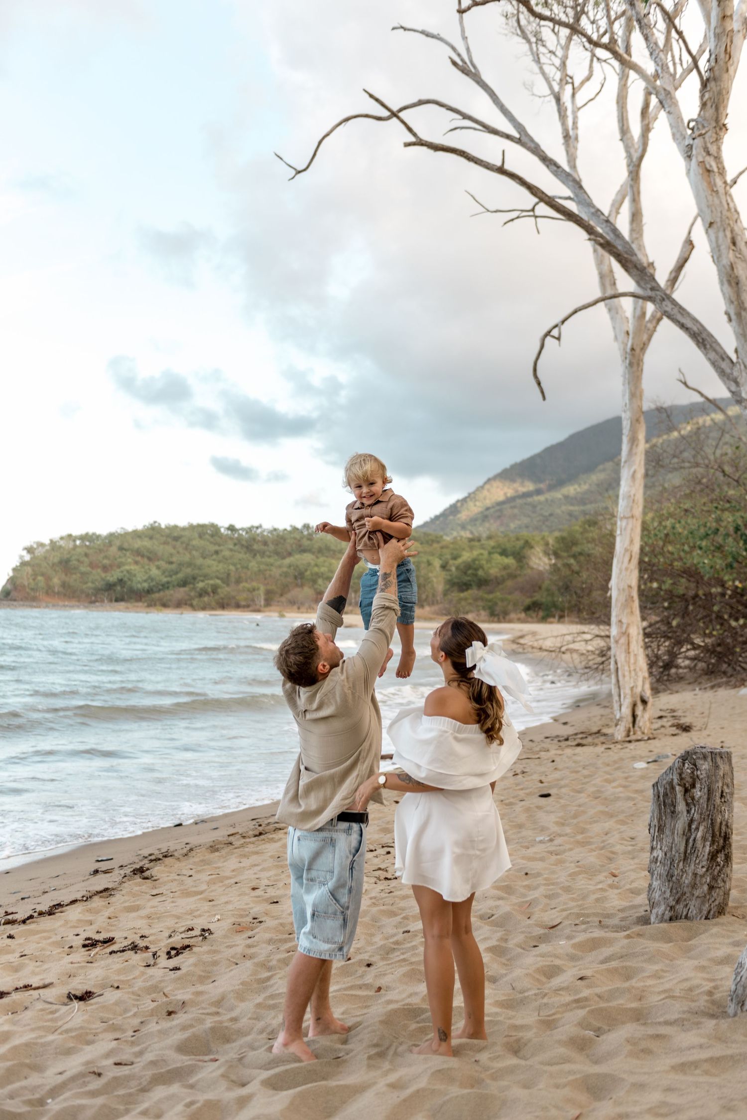 Father tossing toddler in the air on sandy beach while mother watches with white trees nearby.