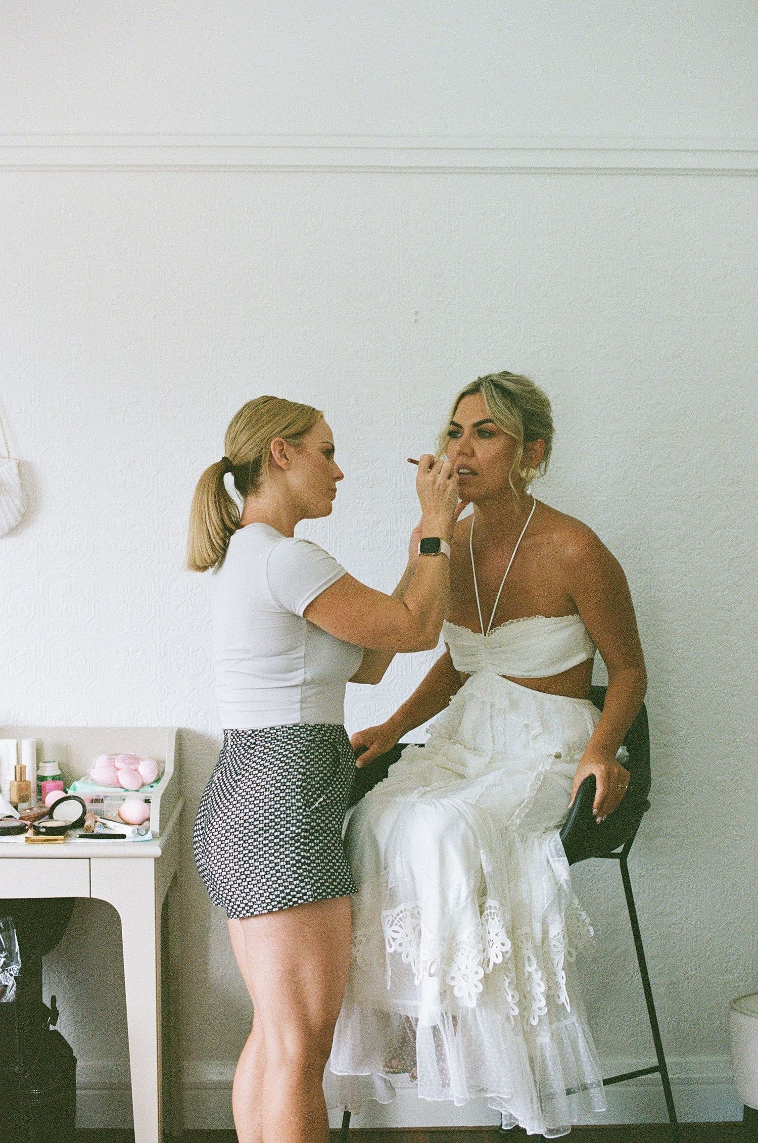 Makeup artist applying lipstick to seated bride in white strapless wedding dress in bright dressing room.