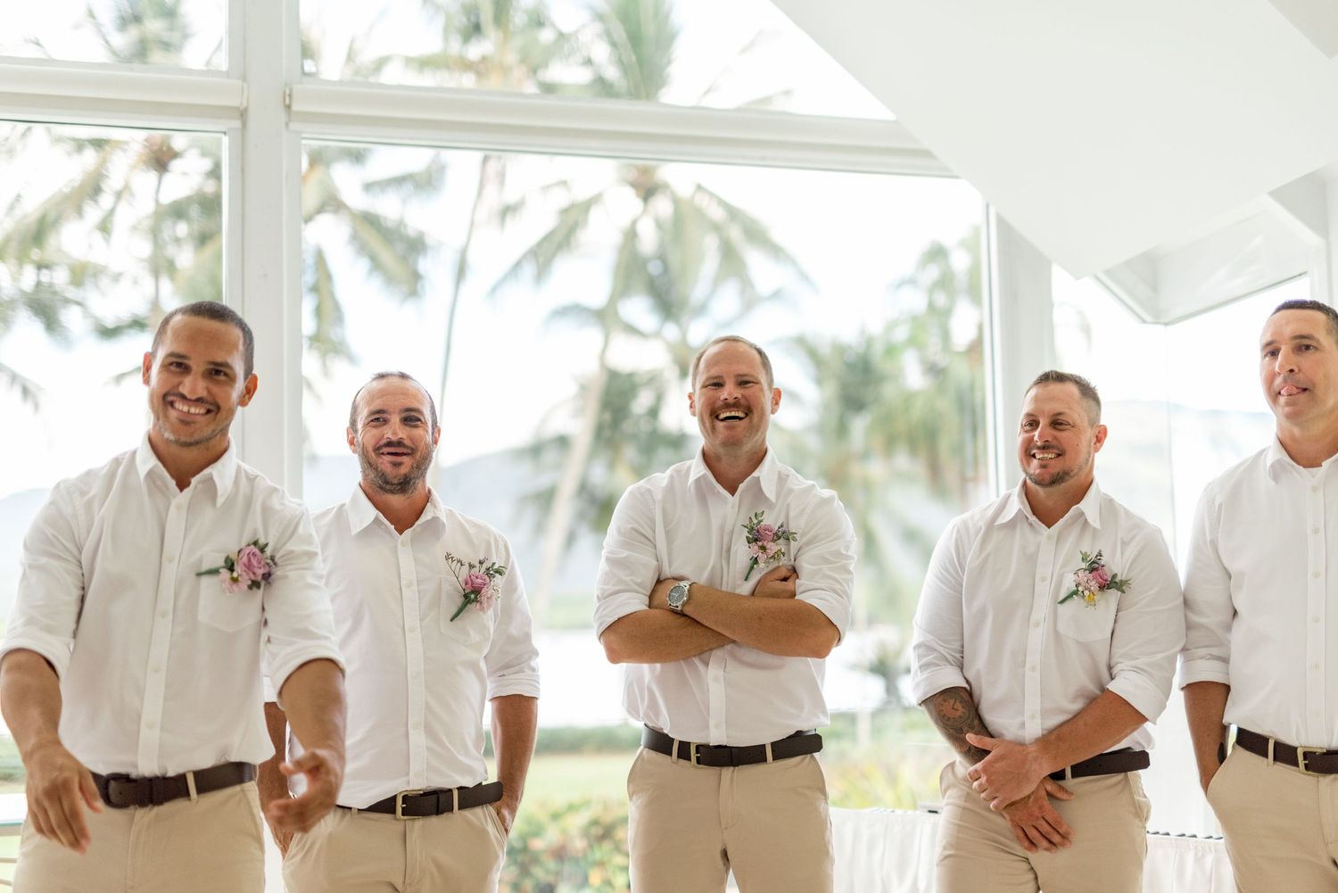 Five groomsmen in white shirts and khaki pants laughing together at a tropical wedding venue.