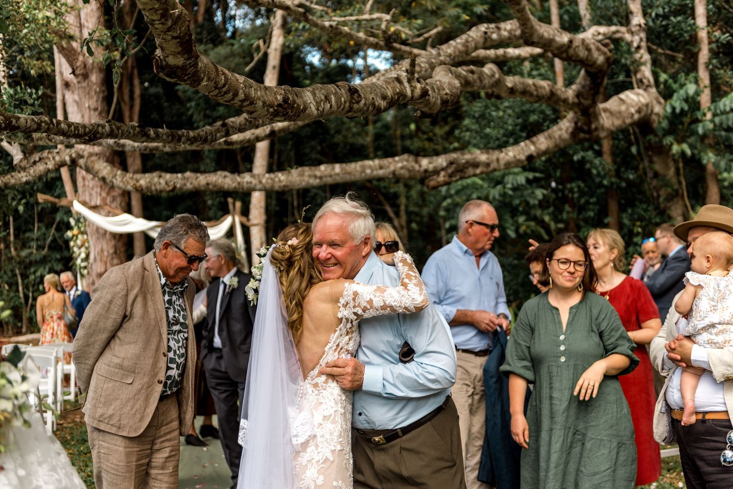 Bride embracing older gentleman guest after outdoor woodland wedding ceremony.