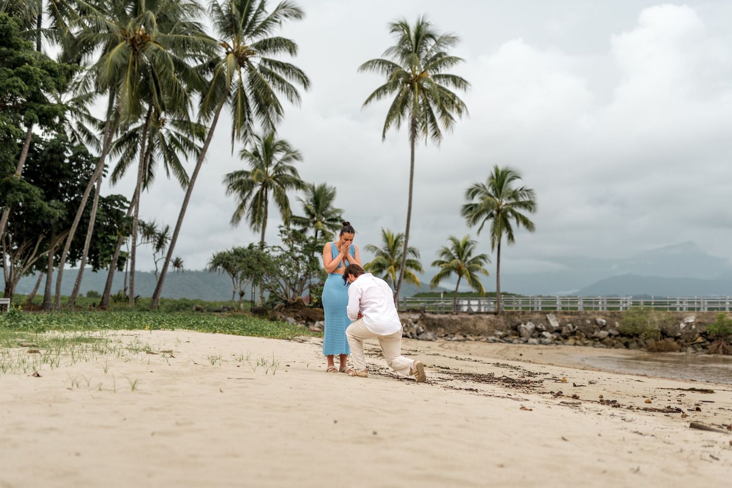 Couple sharing romantic proposal moment on sandy beach surrounded by tall palm trees.