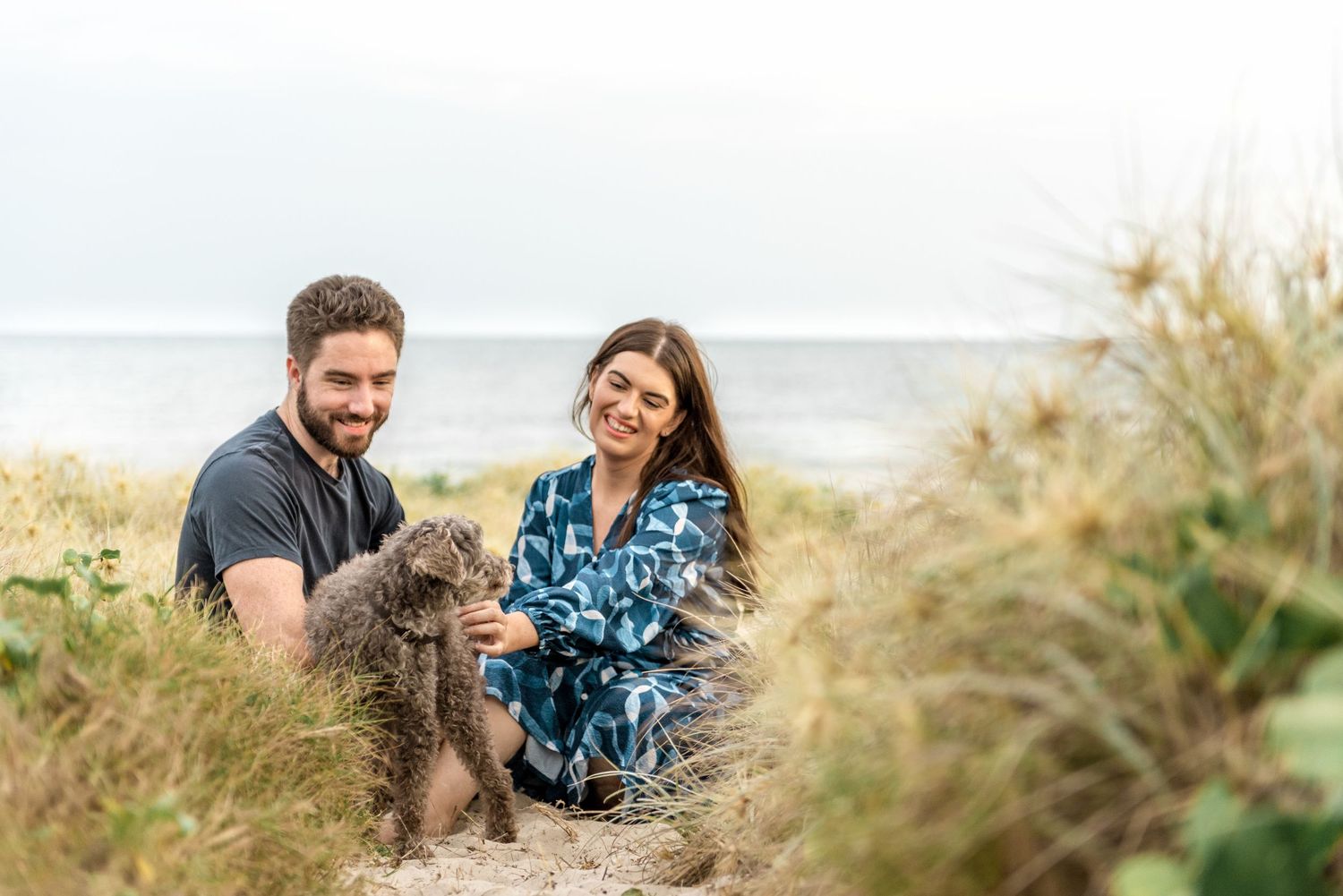 Smiling couple crouching in beach grass dunes with fluffy grey dog by the sea.