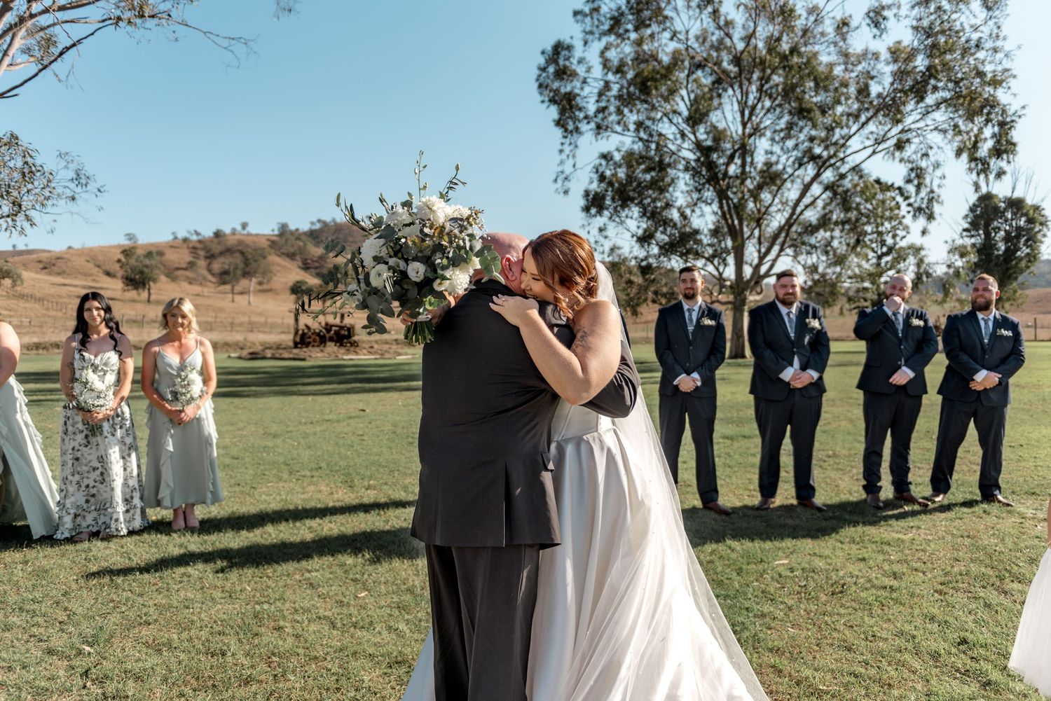 Bride and groom share their first kiss at an outdoor wedding ceremony on a green lawn with bridal party standing behind.