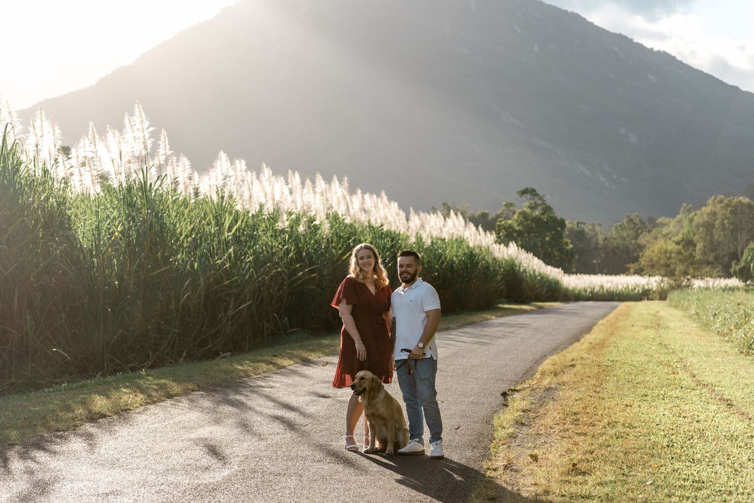 Happy couple walking their golden retriever dog along a peaceful path with mountain and tall grasses behind.