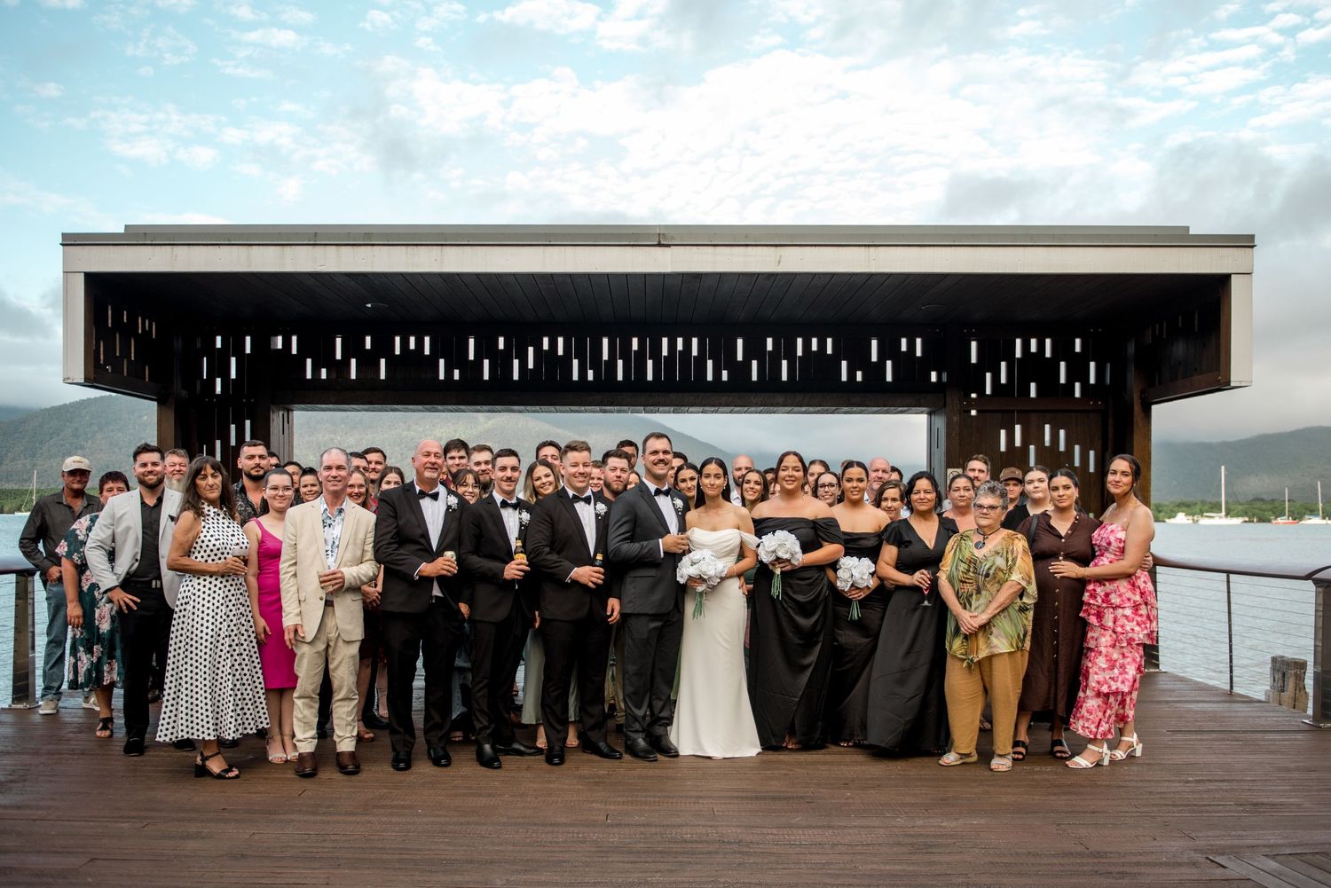 Large wedding party group photo on a waterfront deck with misty mountains in the background.