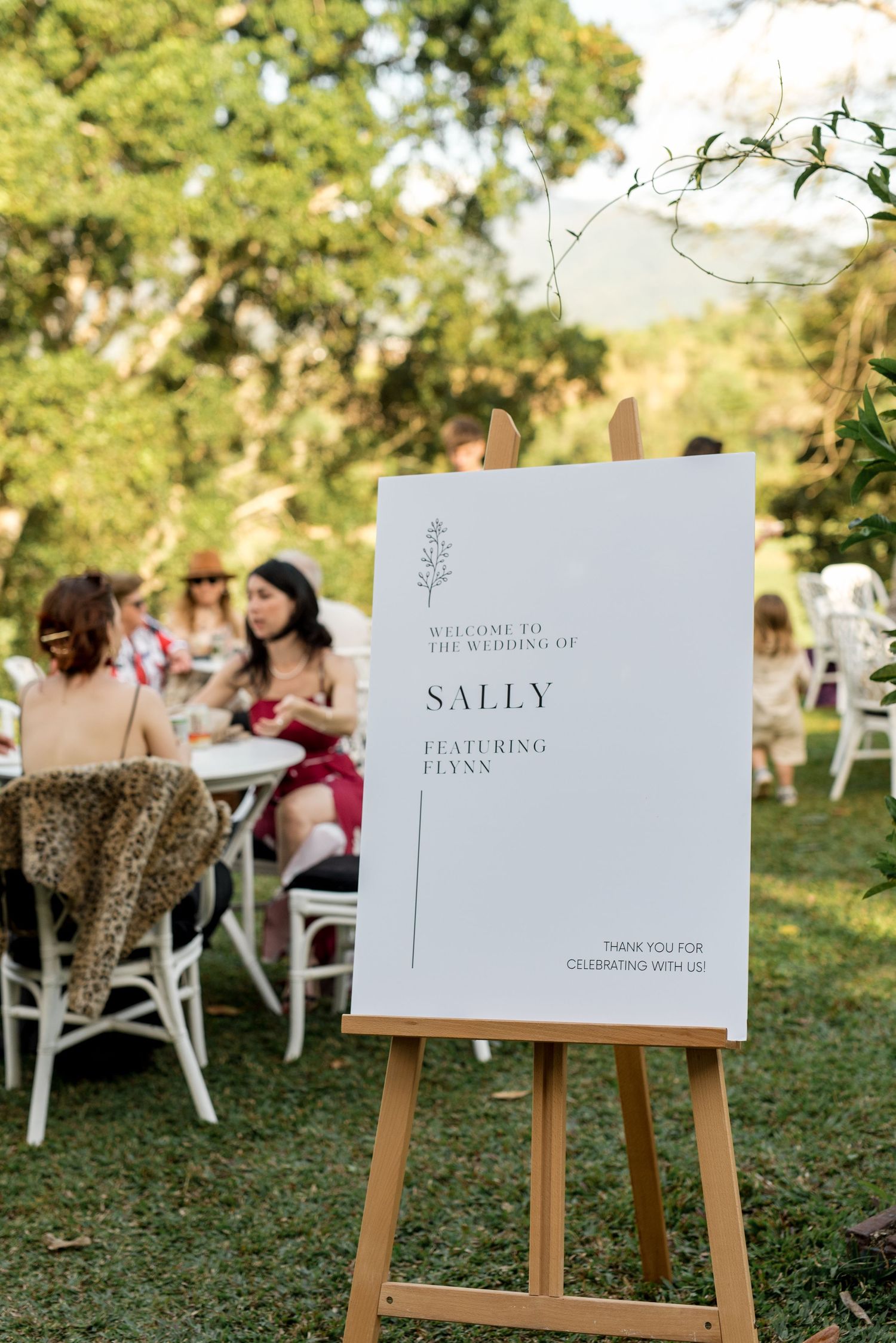 Elegant outdoor wedding welcome sign for Sally and Flynn displayed on wooden easel among garden guests.