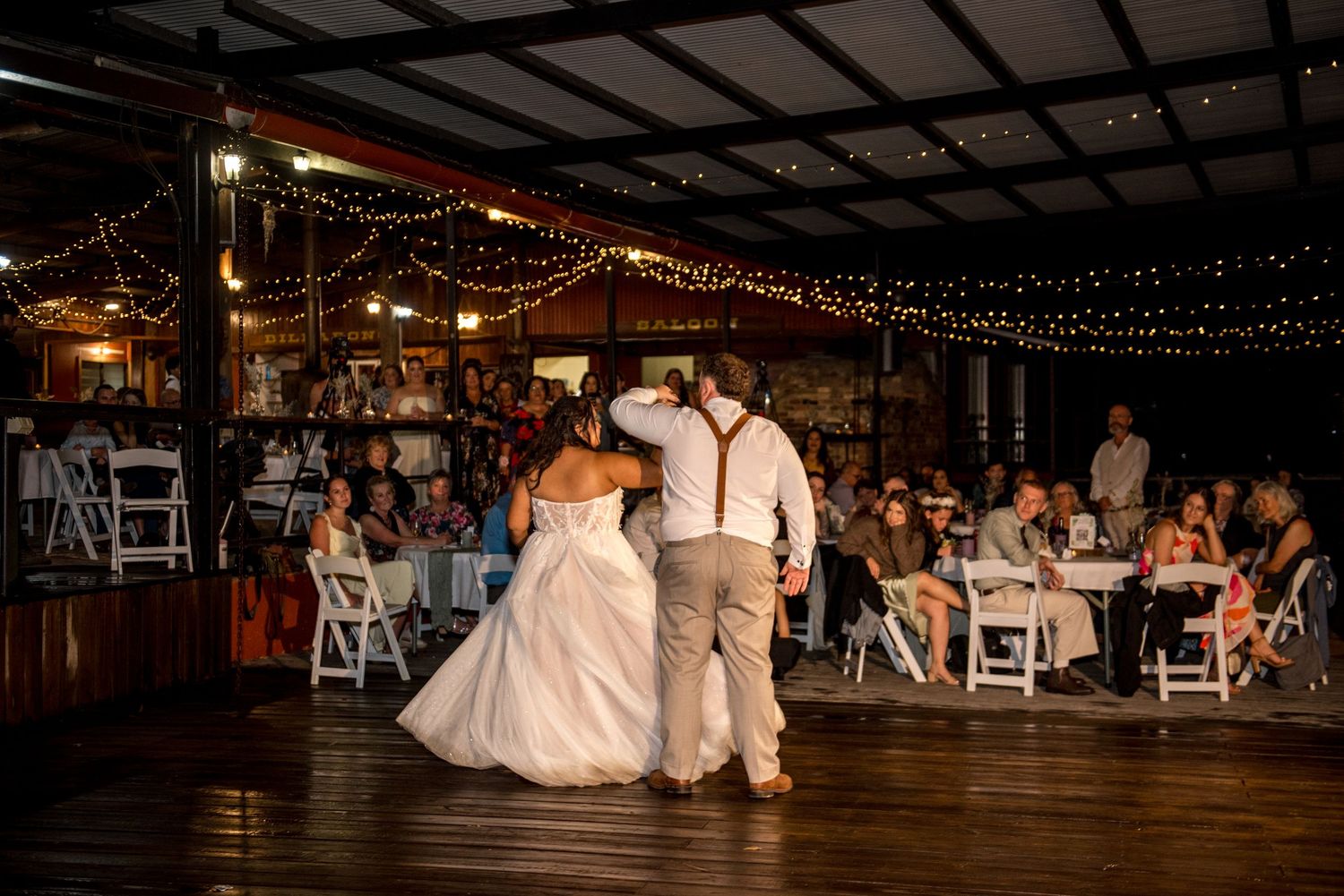 Bride and groom share first dance on wooden floor with wedding guests watching under string lights.