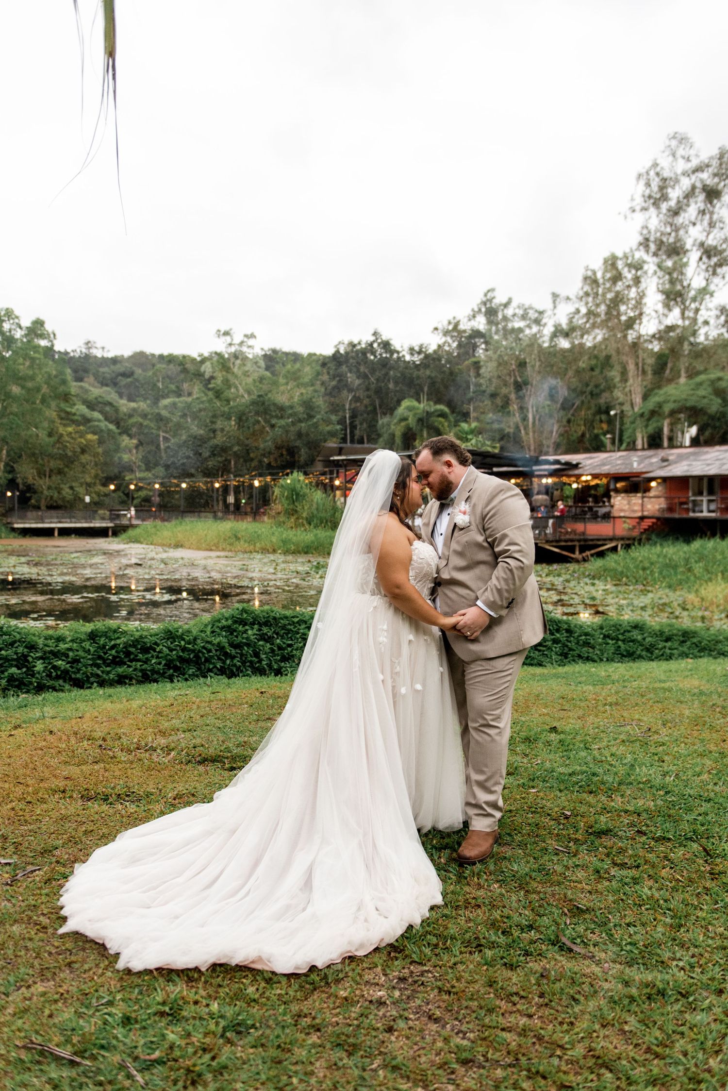 Bride and groom share an intimate embrace on lush green lawn with a scenic pond backdrop.