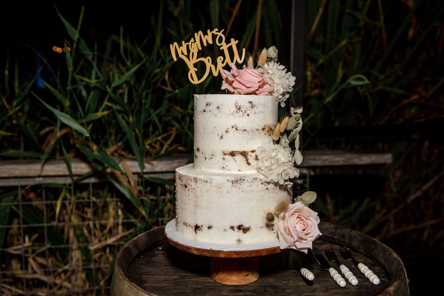 Elegant two-tier naked wedding cake with gold topper, pink roses, and white florals on wooden barrel.