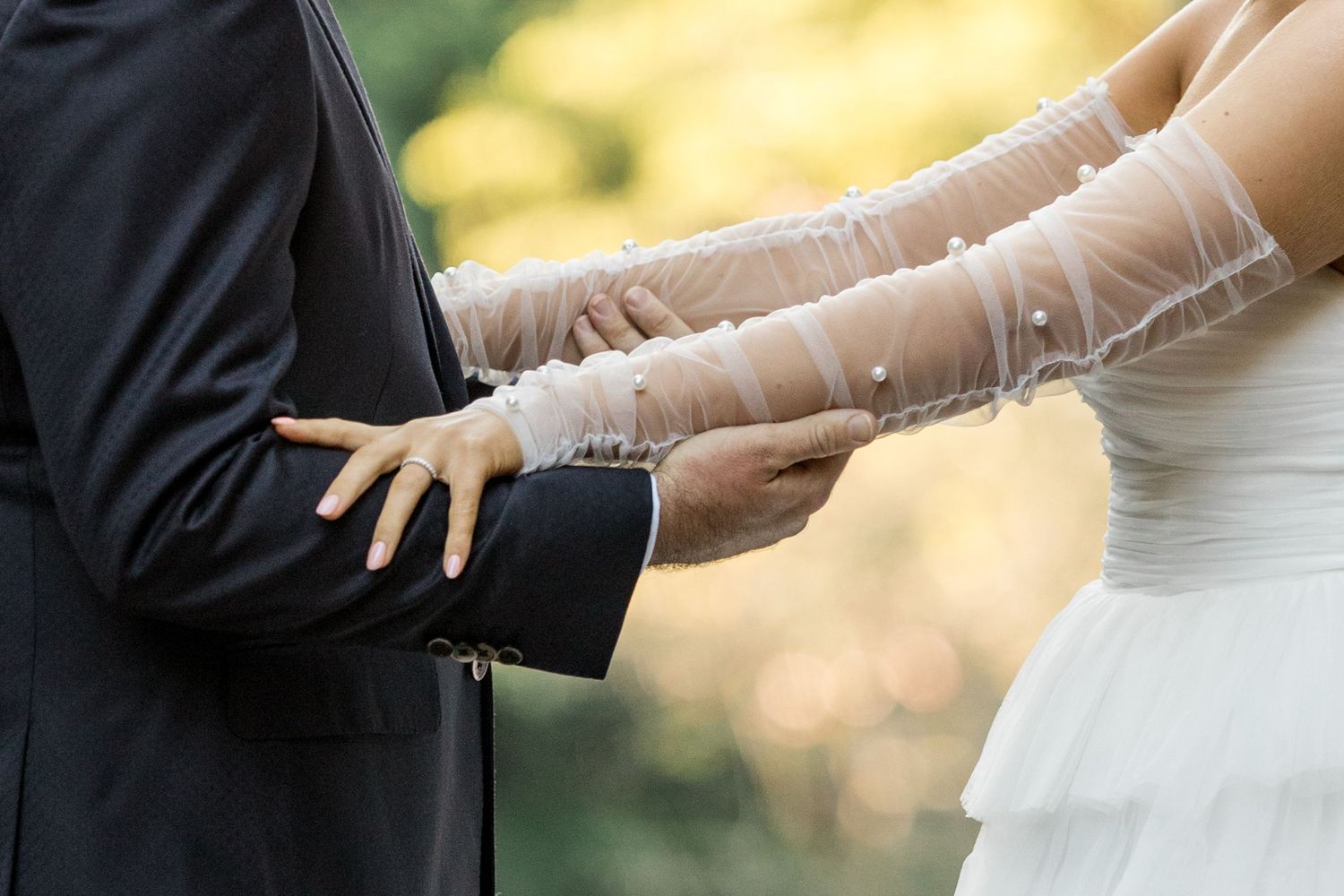 Bride and groom hold hands during wedding ceremony, bride wearing elegant lace gloves and white gown.