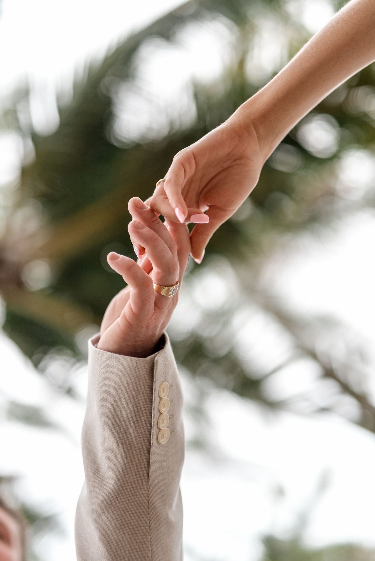 Close-up of bride and groom gently touching fingertips with gold wedding bands against blurred palm trees.