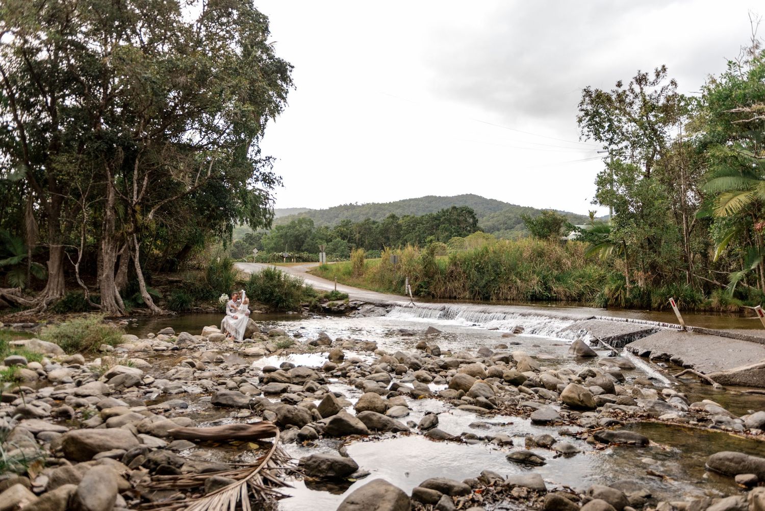 Scenic rocky creek landscape with bride and groom visible among lush green mountain scenery.