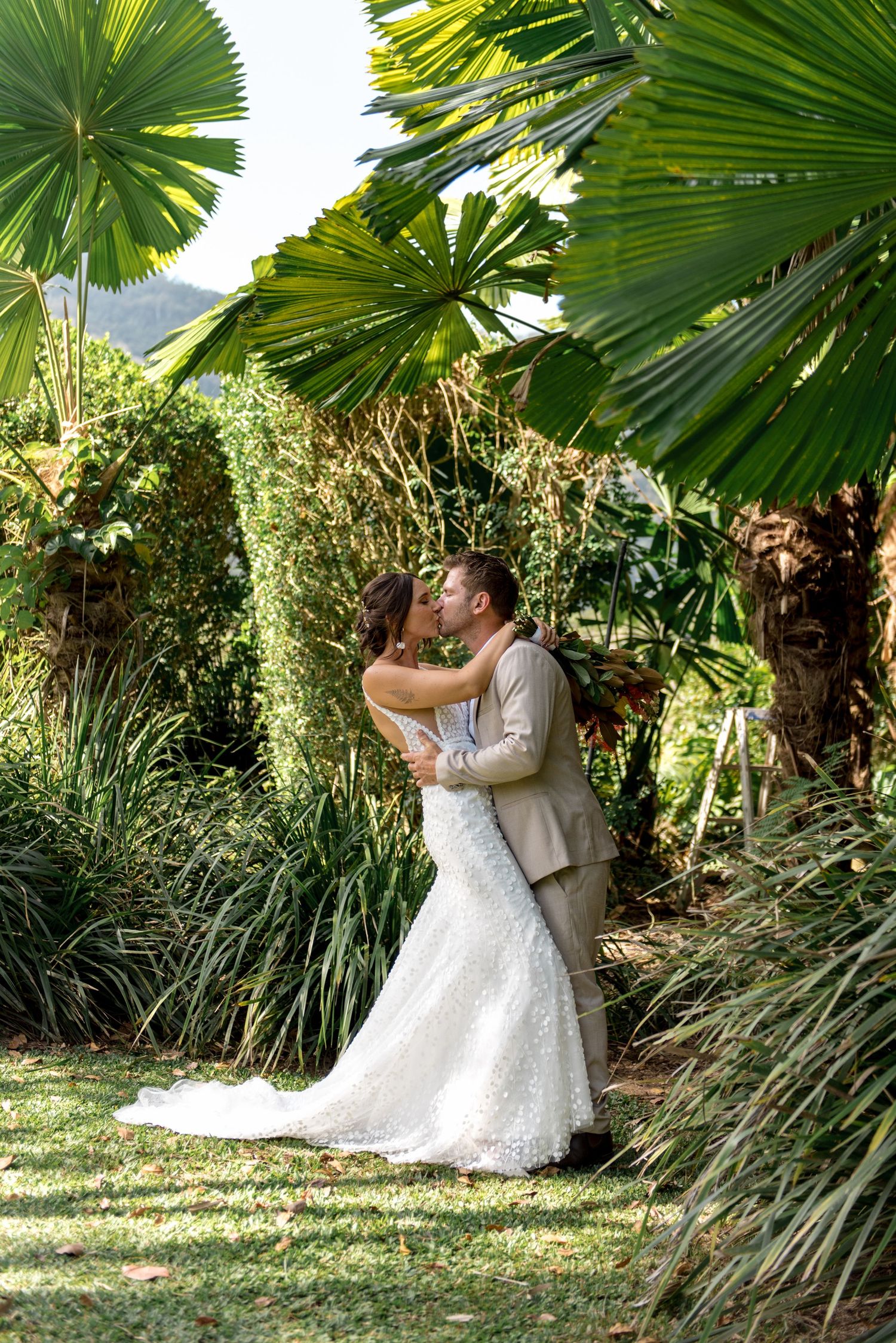 Wedding couple embracing and kissing surrounded by vibrant green tropical fan palms outdoors.