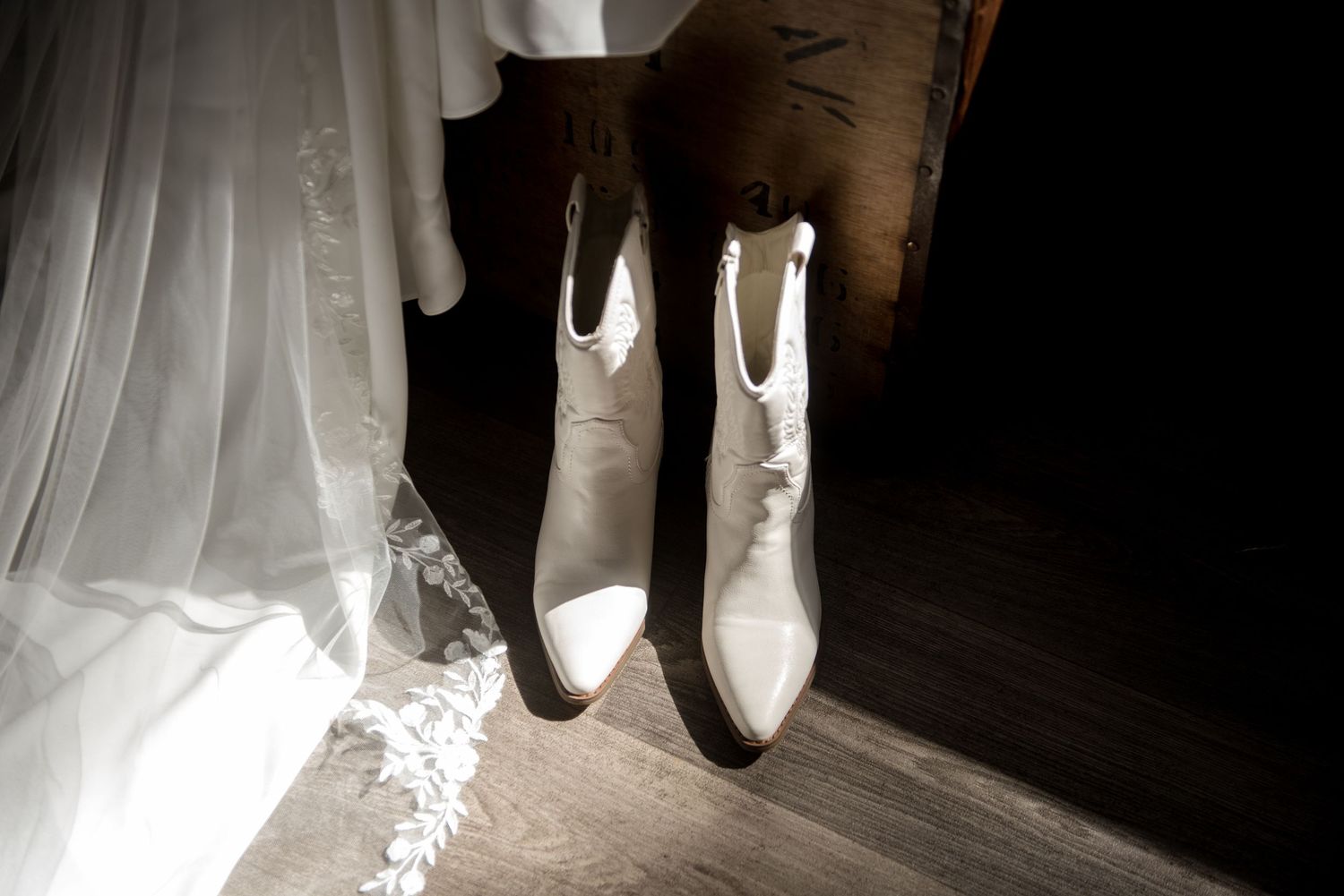 White cowboy bridal boots beside a flowing lace wedding dress on wooden floor.