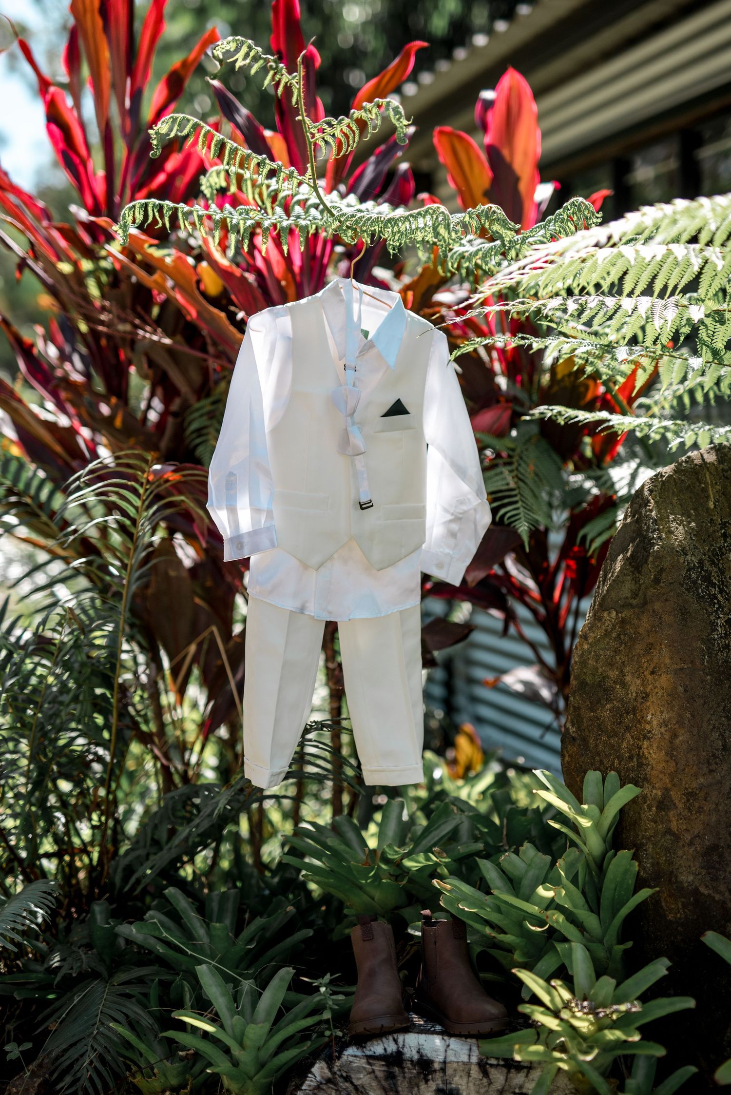 Small white ring bearer suit displayed hanging on tropical red foliage in garden setting.