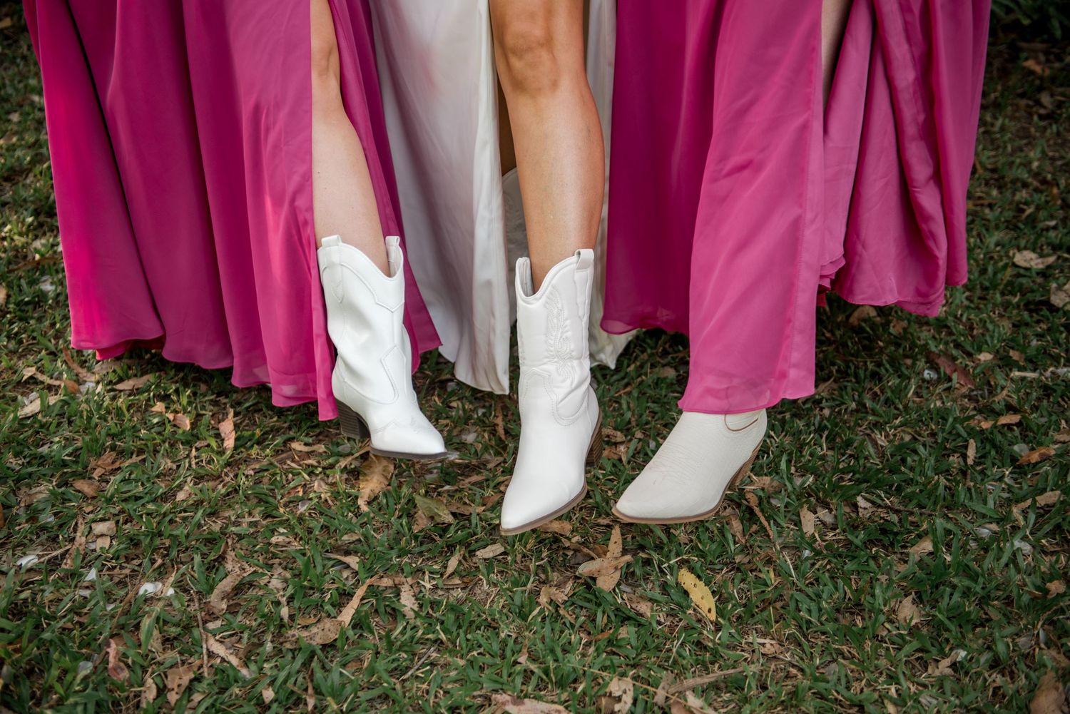 Bride wearing white cowboy boots stands between bridesmaids in pink dresses on grass at wedding.