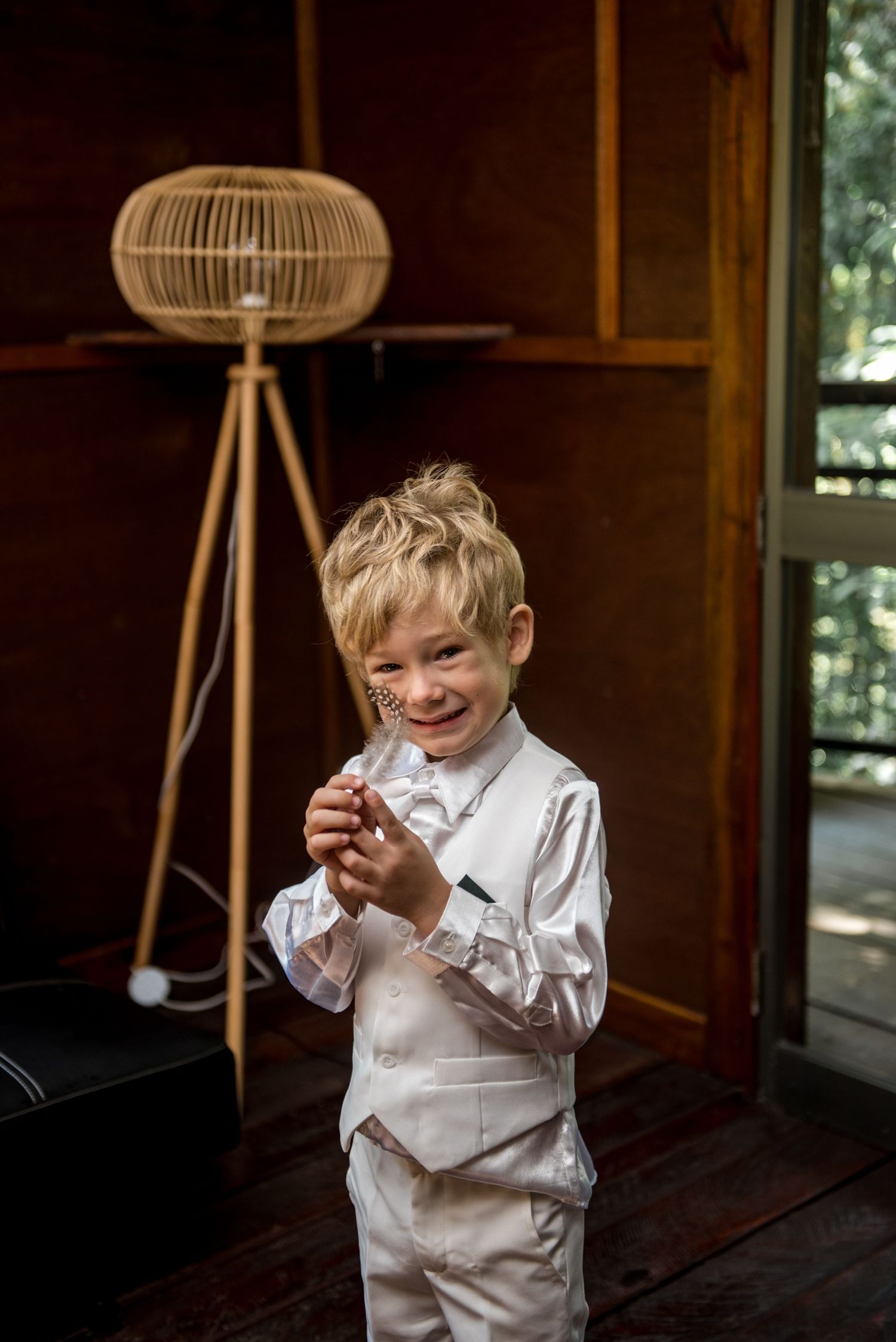 Young boy in white suit smiling while holding item in rustic wooden room.