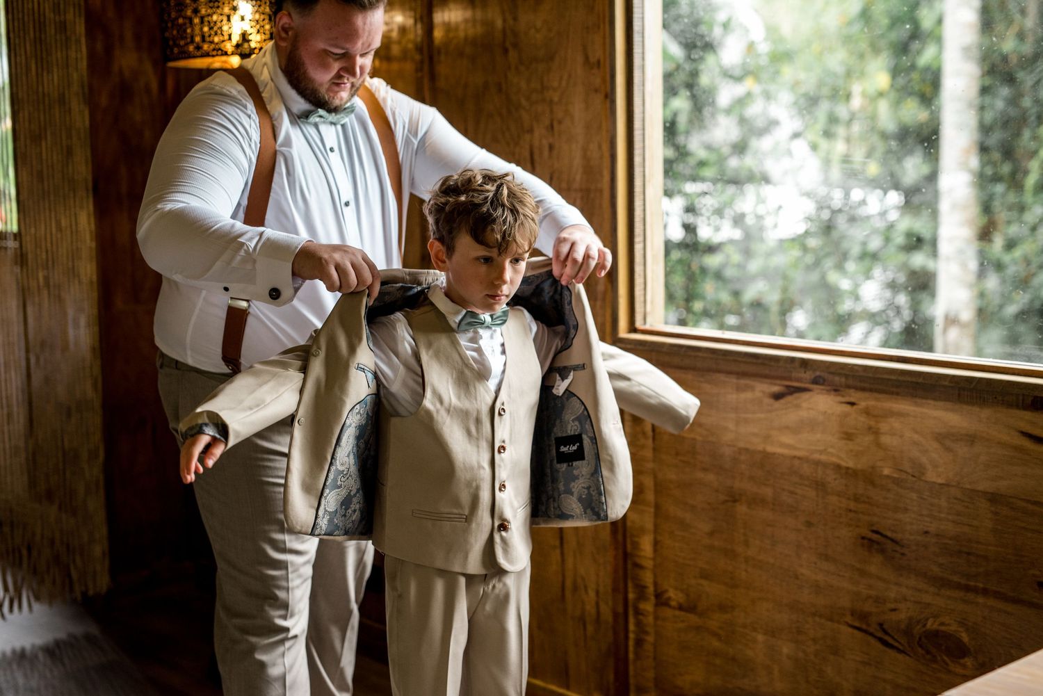 Groom in suspenders helping young ring bearer put on his beige suit jacket near a window.