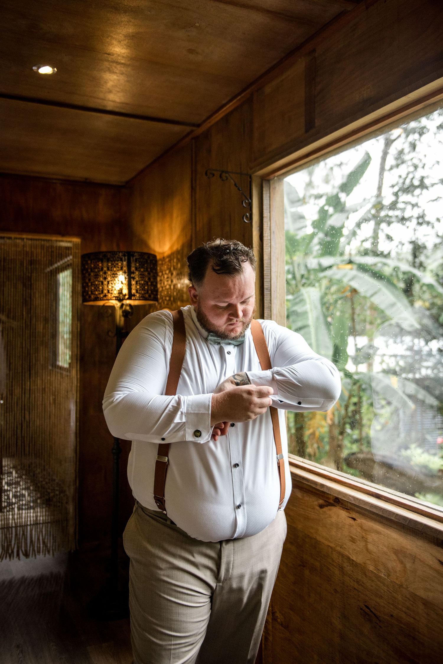 Groom in white shirt and brown suspenders checking his watch while getting ready near a wooden cabin window.