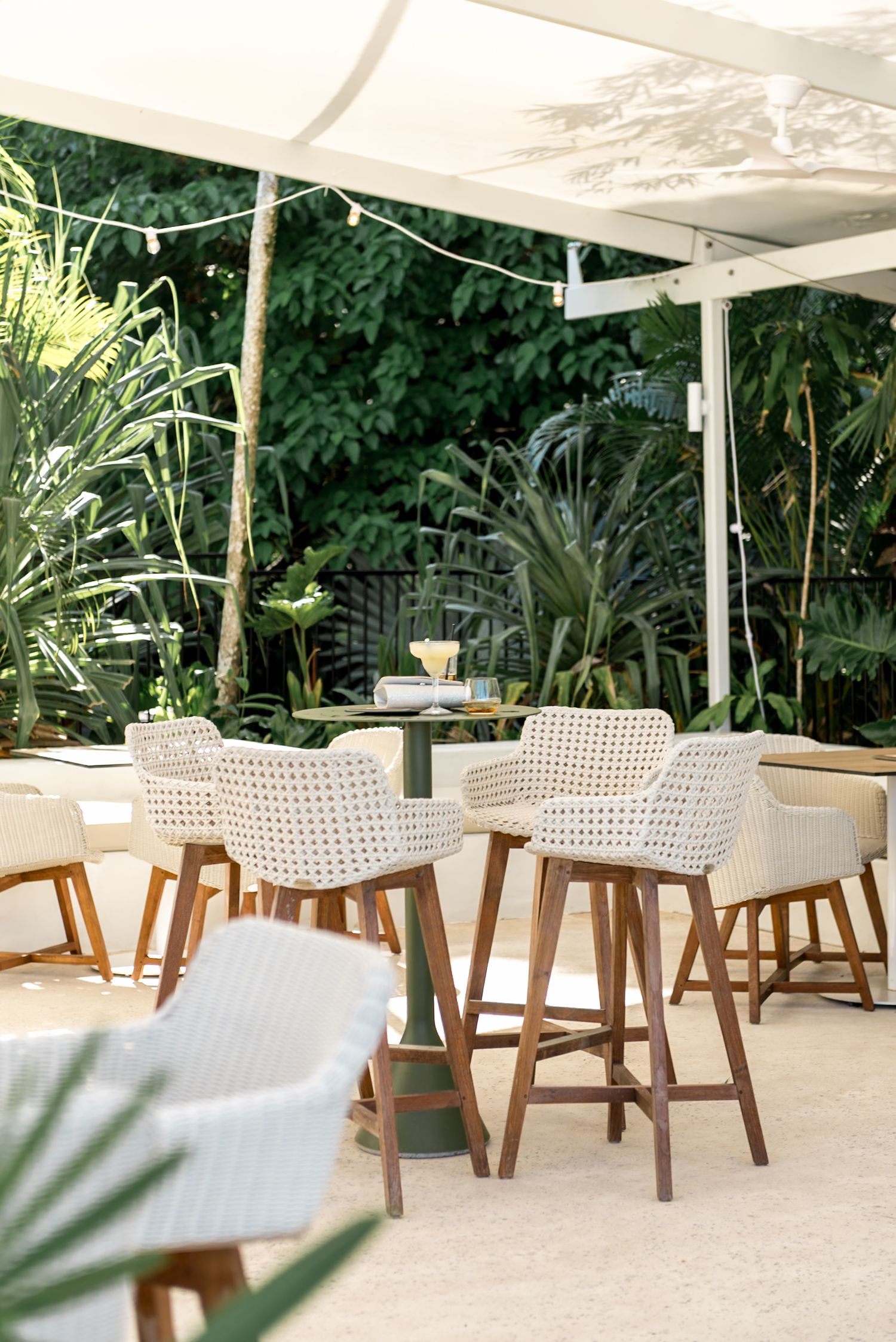 Elegant outdoor wedding reception area with wicker bar stools, string lights, and lush tropical greenery.