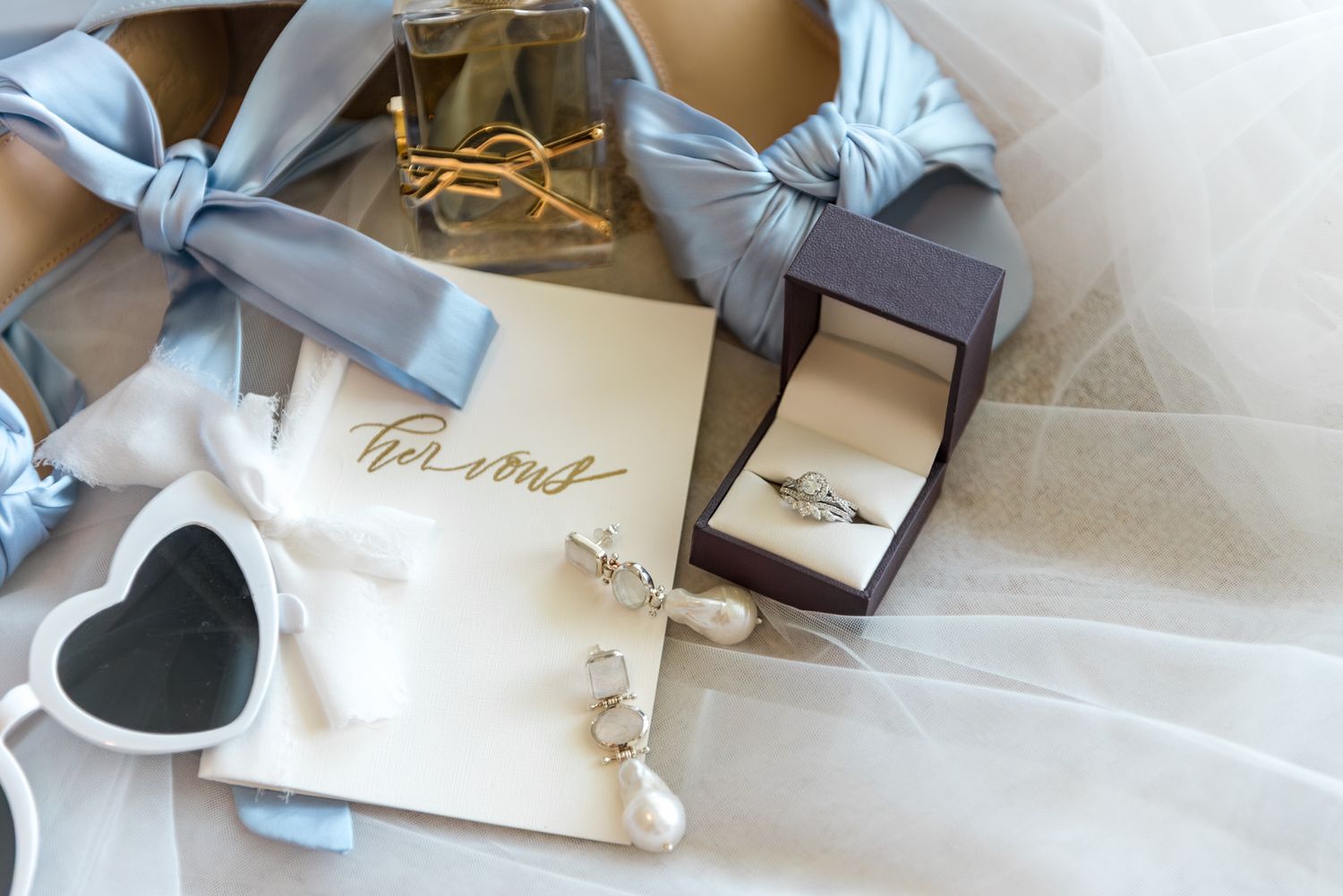 Bridal accessories flat lay with blue heels, diamond ring box, pearl earrings, and YSL perfume on veil.