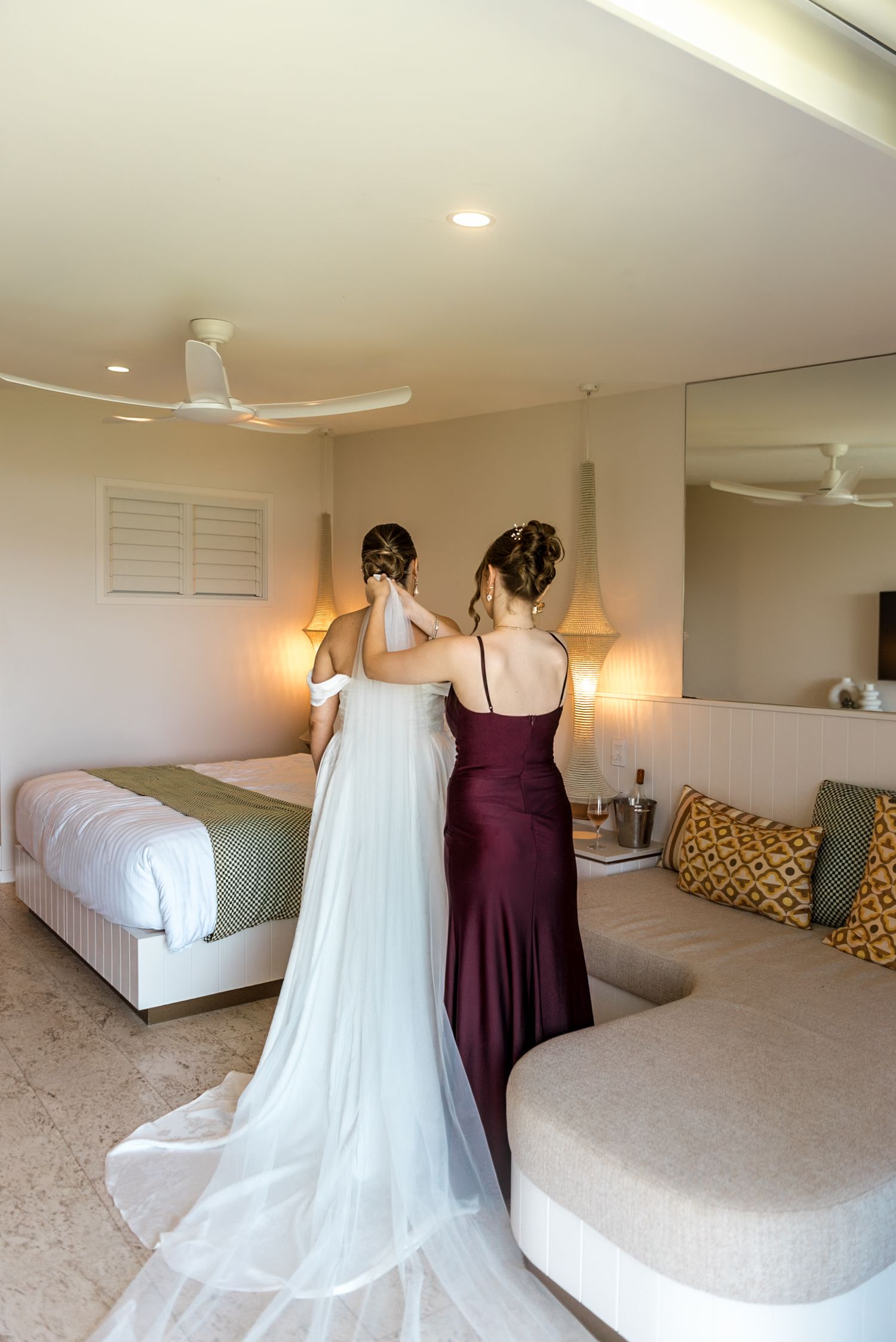Bridesmaid in burgundy gown helping bride in white off-shoulder dress with veil in a luxury hotel room.