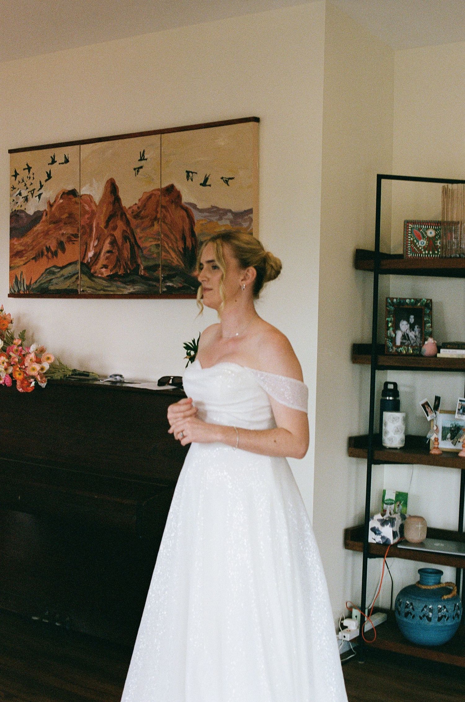Bride in off-shoulder white sparkly gown standing elegantly in a room with mountain artwork.