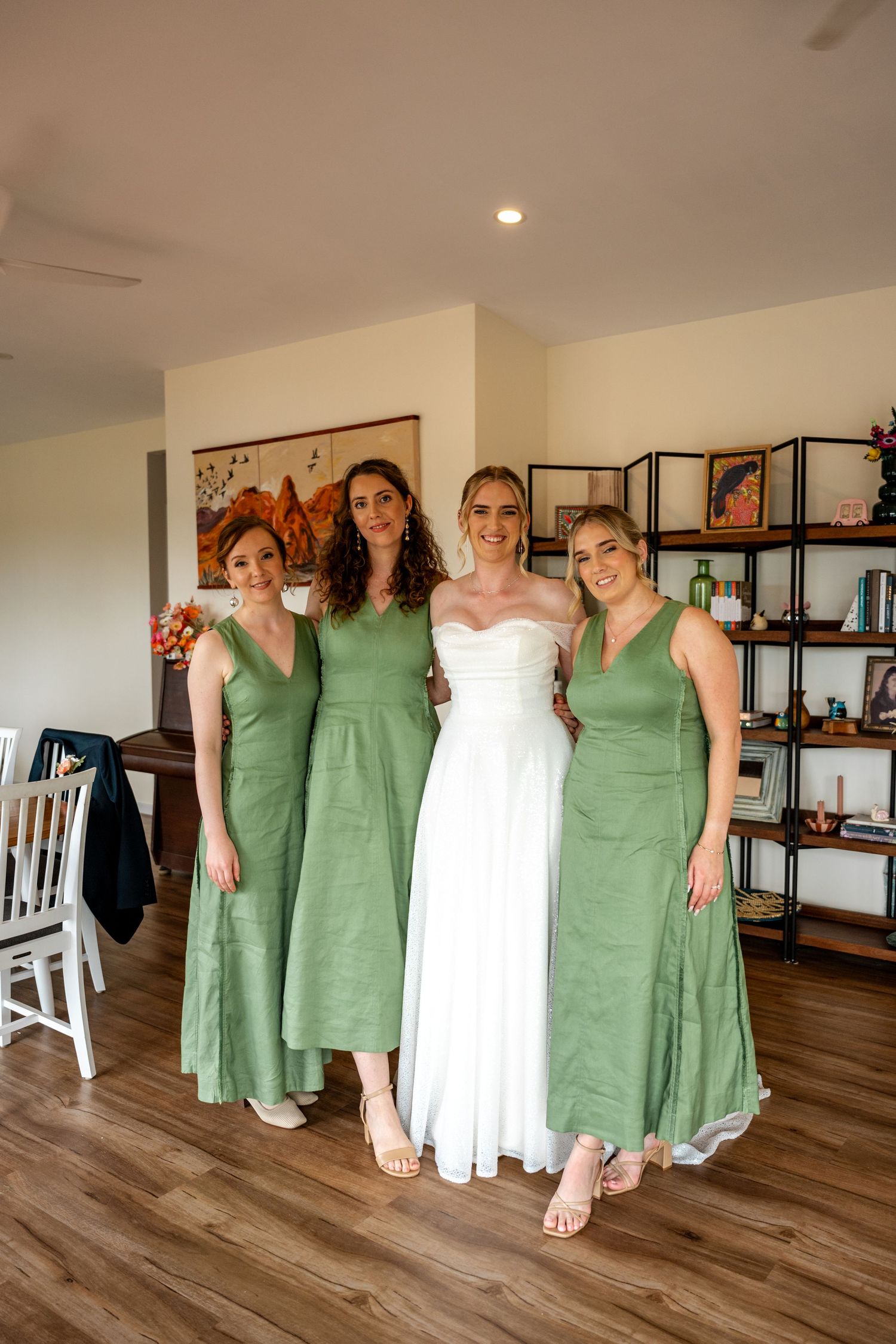 Bride in white gown posing with three bridesmaids in matching sage green dresses indoors.