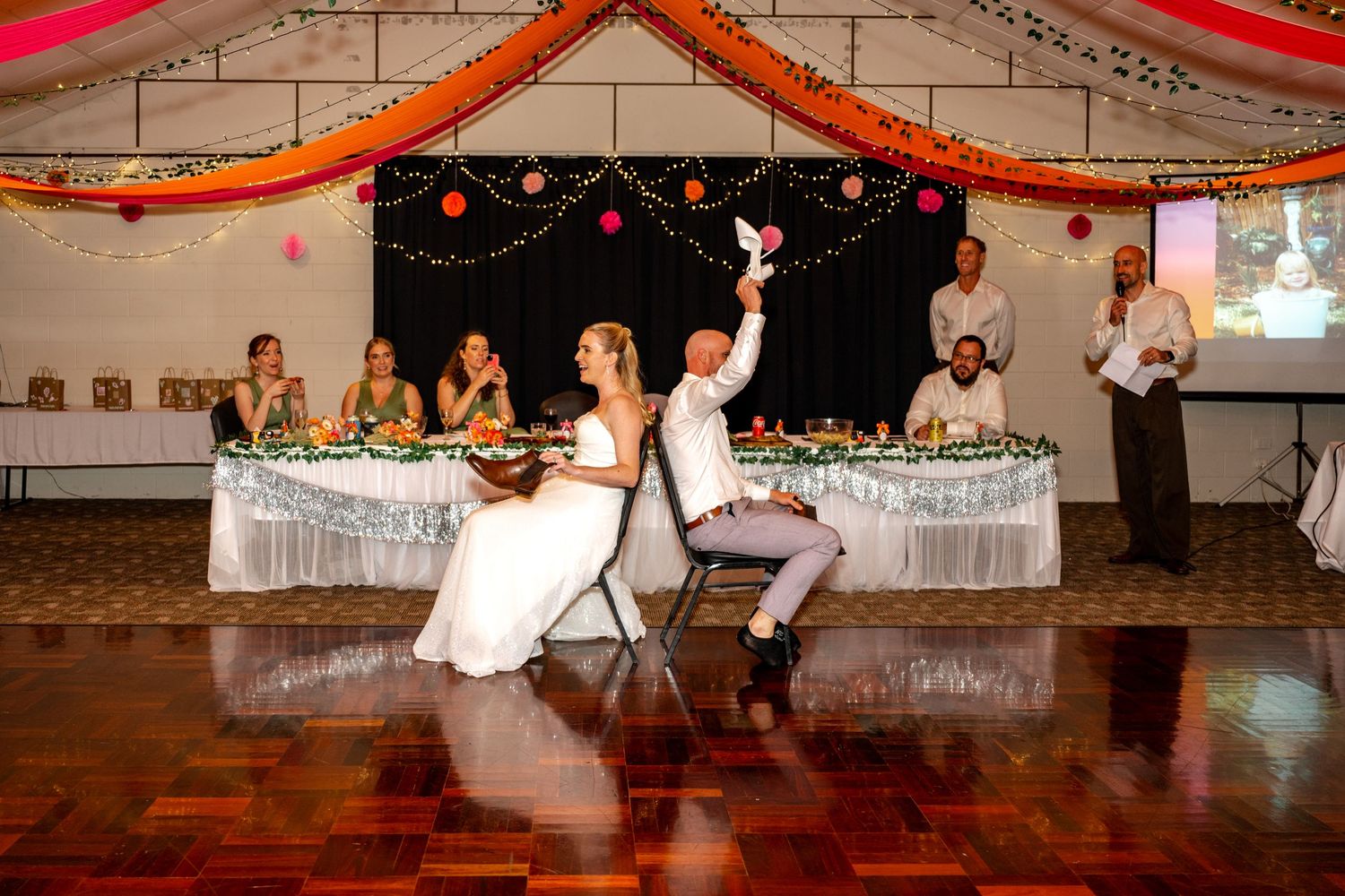 Bride and groom perform fun wedding reception game on dance floor beneath colorful hanging decorations in banquet hall.