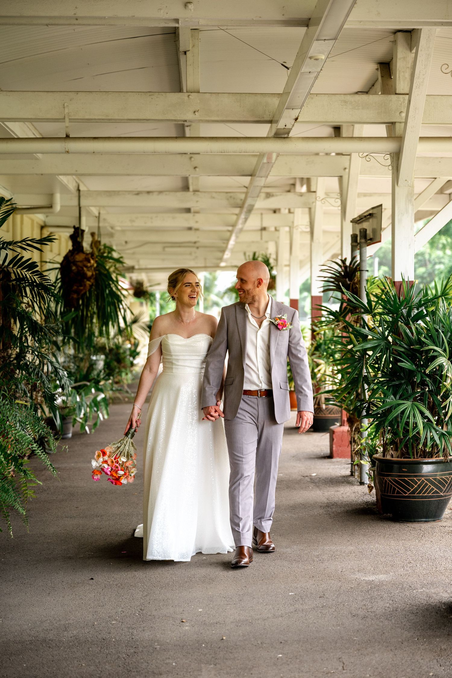 Bride and groom walk hand-in-hand through a covered tropical venue corridor lined with potted plants.
