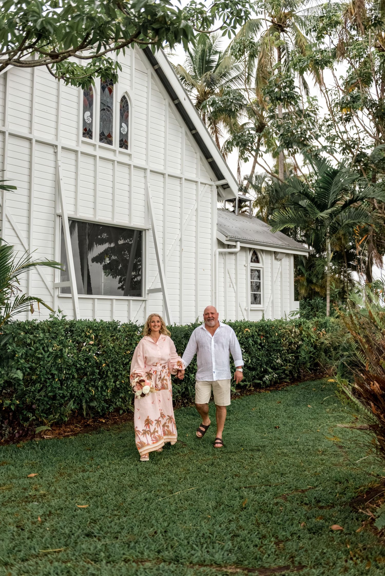 Couple strolling hand in hand across green lawn beside a white timber heritage chapel.