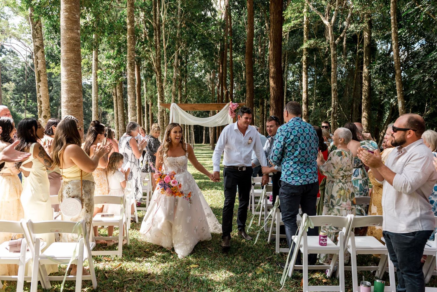 Smiling newlyweds walk hand in hand down the aisle as guests applaud during outdoor forest wedding reception.