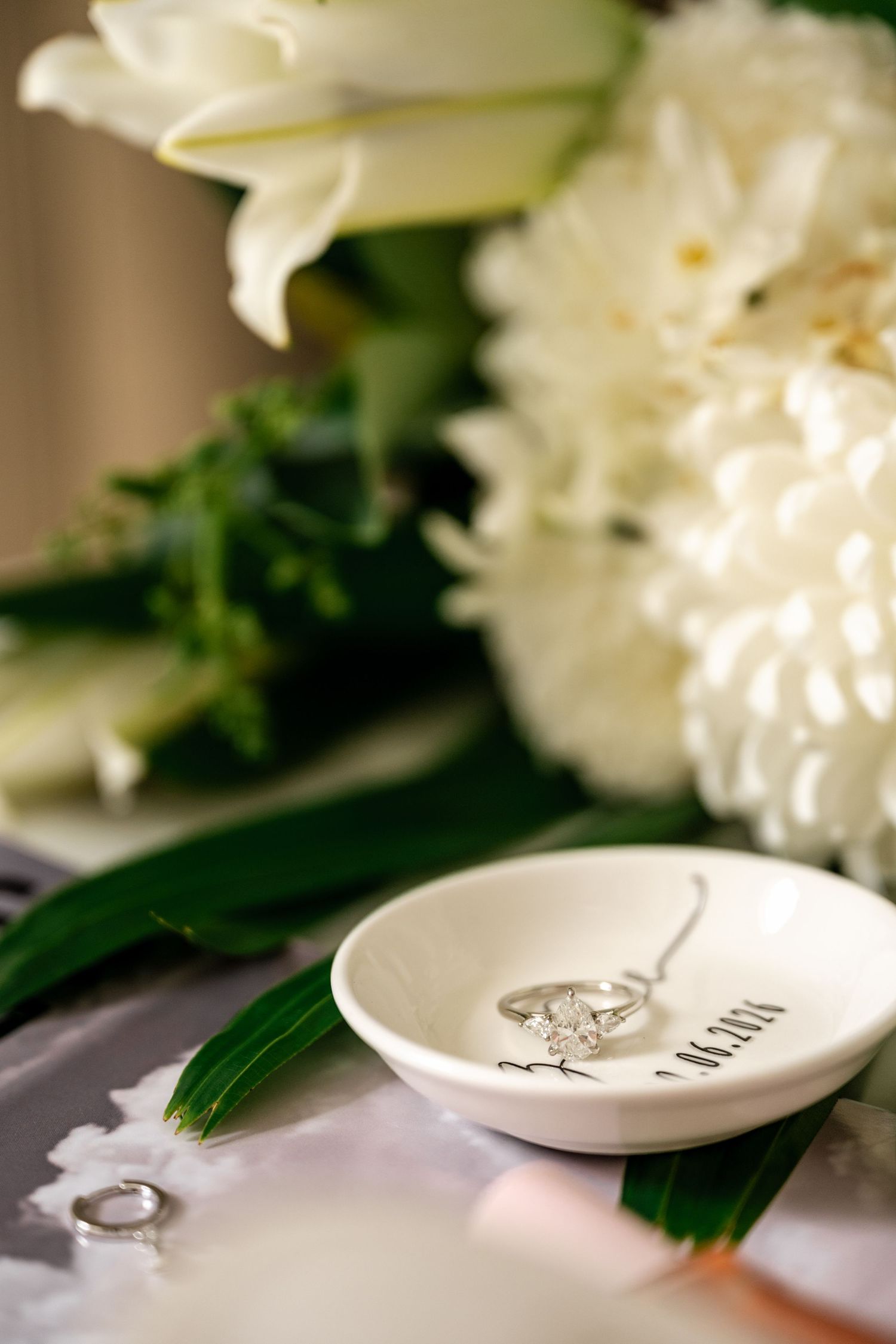 Diamond engagement ring in a personalized white ceramic dish beside a white floral bouquet.