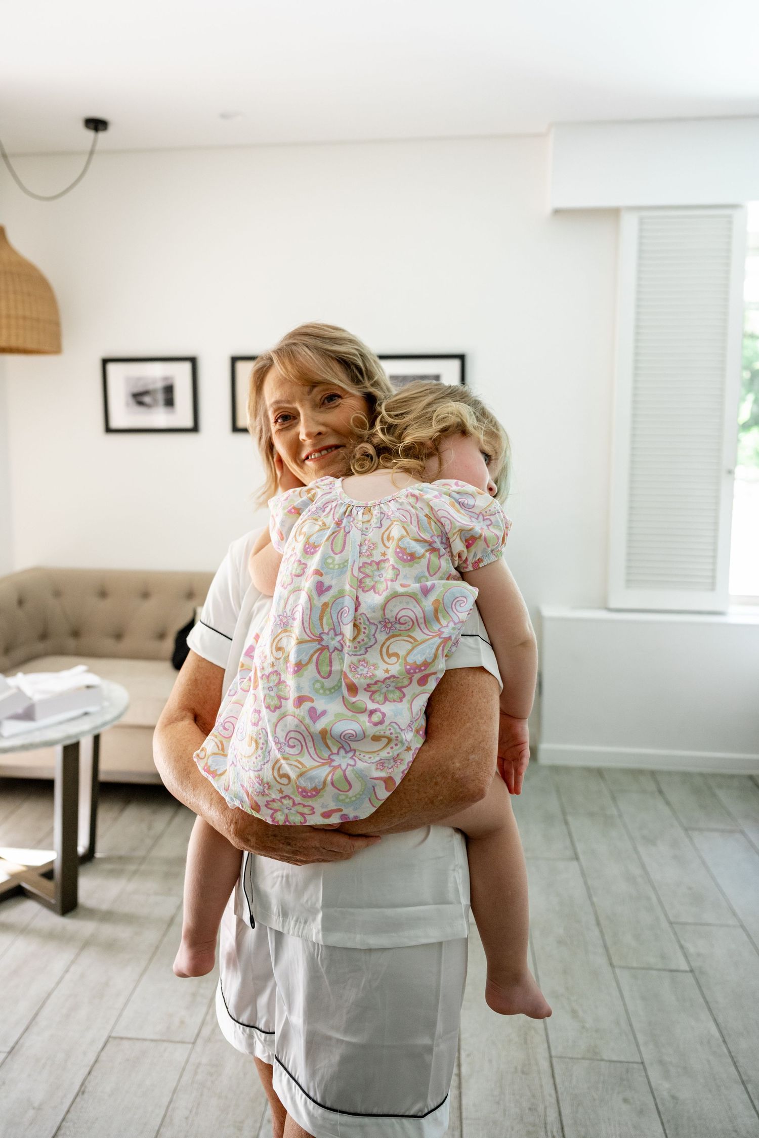 Smiling blonde woman holding a toddler girl in a floral outfit in a bright modern living room.