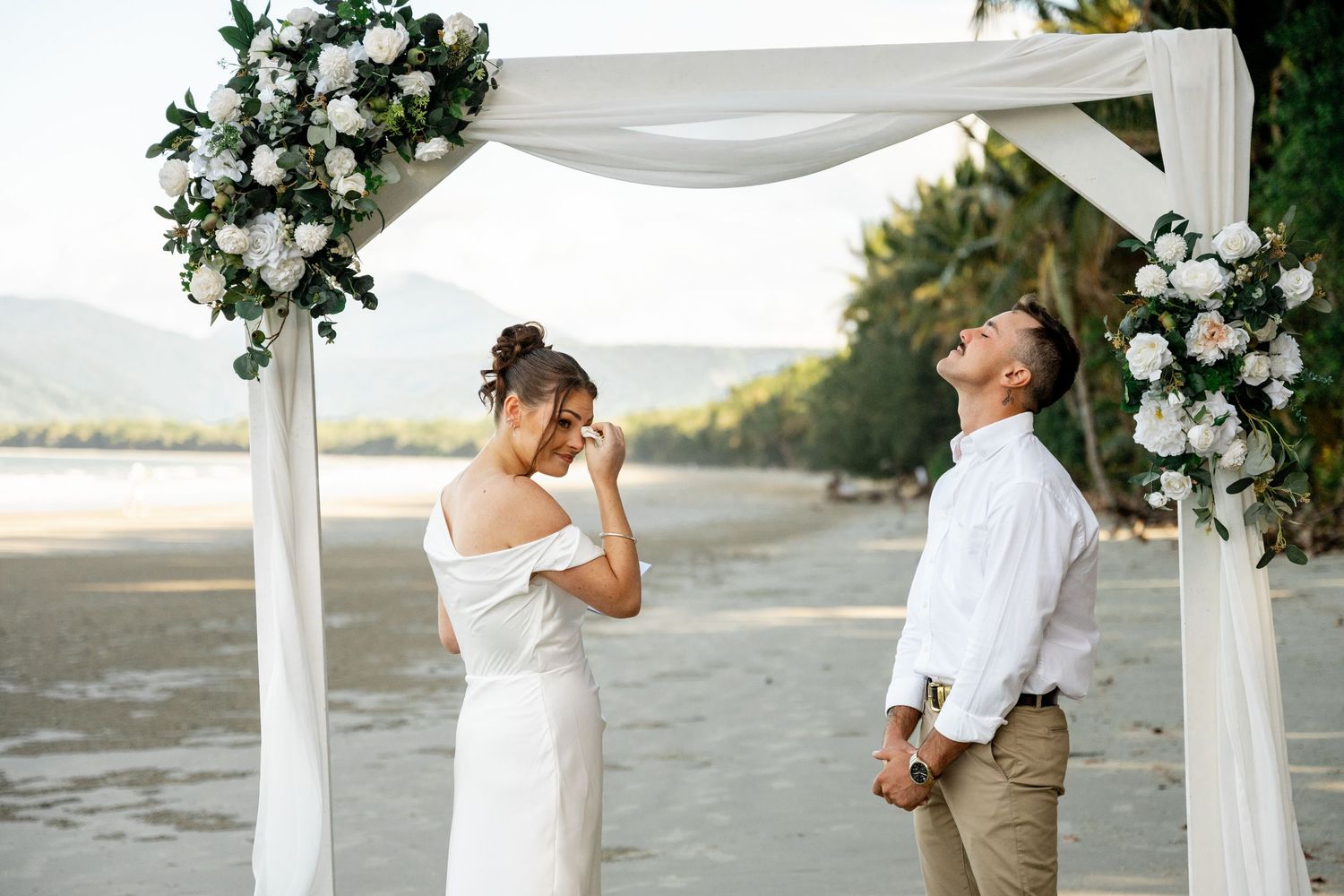 Emotional bride and laughing groom sharing a joyful moment under a white floral wedding arch on the beach at Four Mile Beach, Port Douglas.