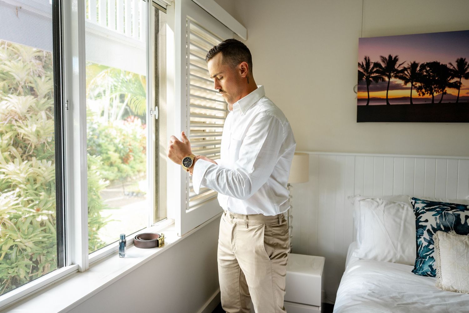 Groom in white shirt and khaki pants looking out a bedroom window on his wedding morning.