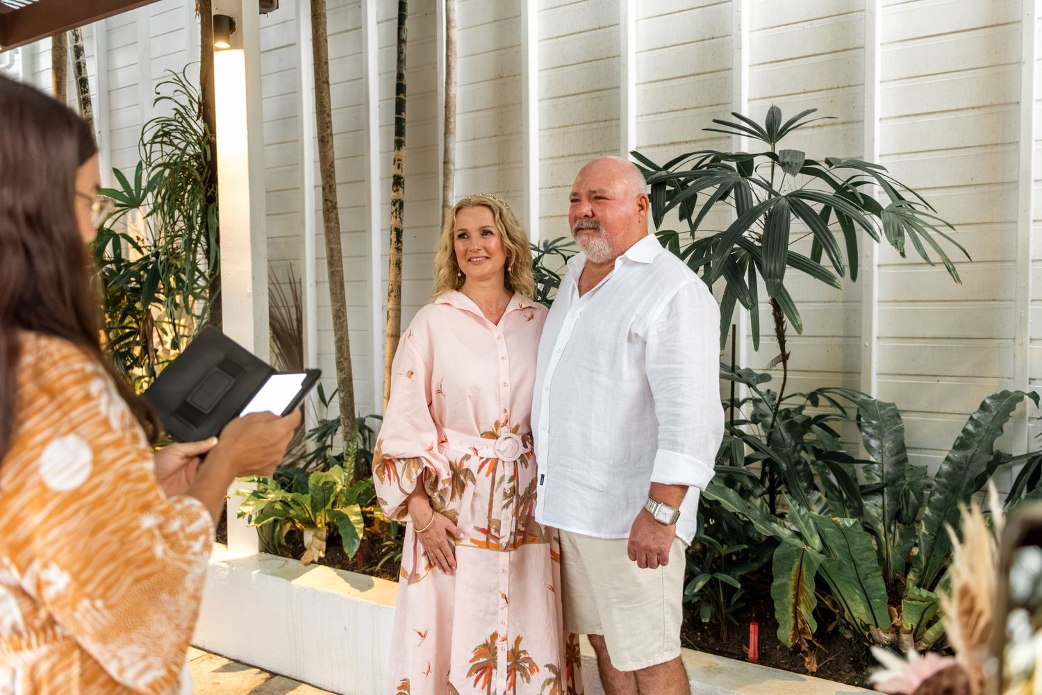 Smiling mature couple standing before tropical plants while a woman reads from a tablet.