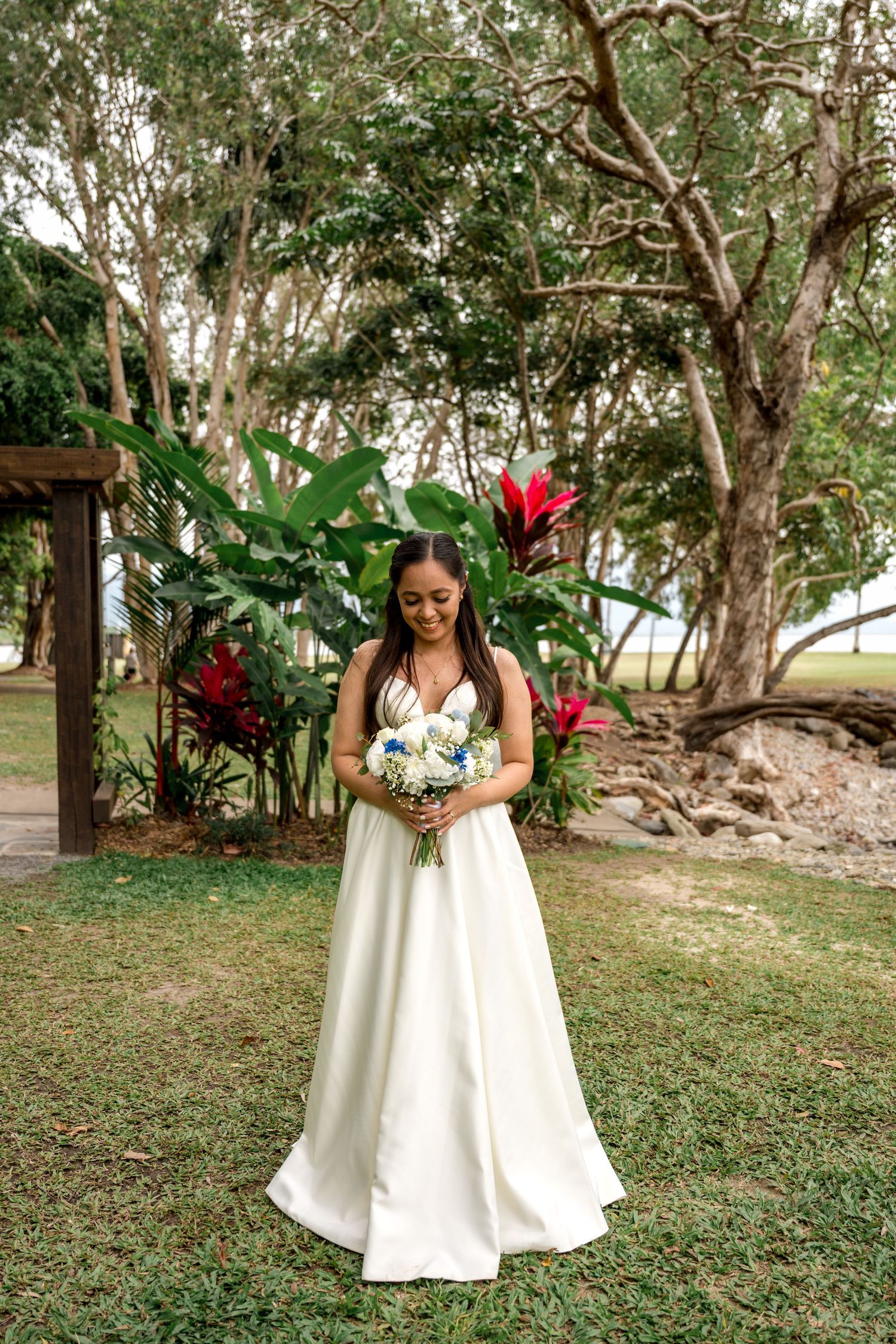 Bride in elegant white satin gown looks down at bouquet in tropical garden setting at Little Cove, Port Douglas