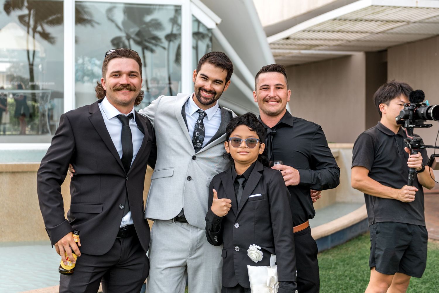 Groom poses with groomsmen and young boy in matching black suits outside modern wedding venue.