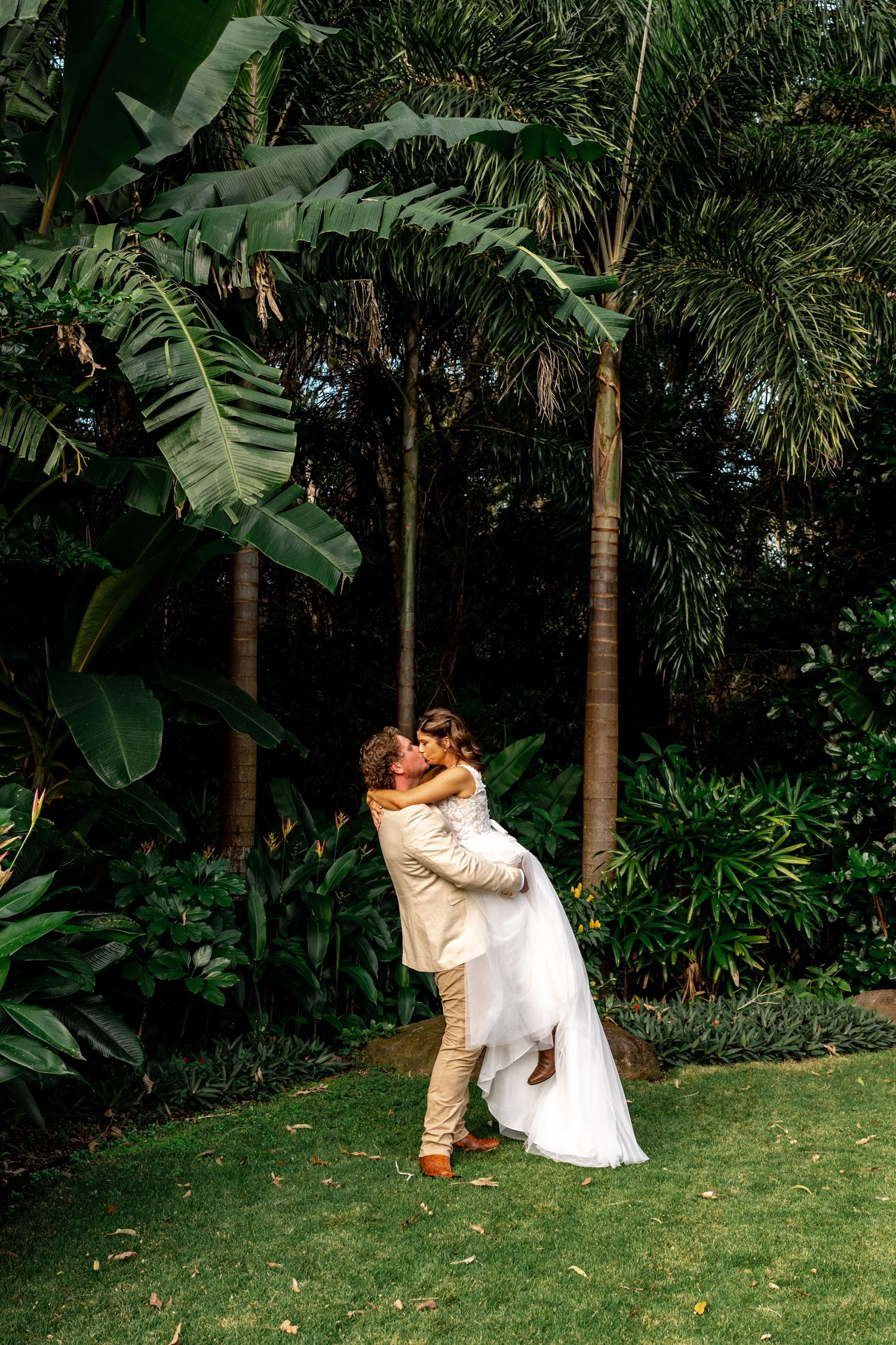 Groom lifting bride in white gown for romantic kiss amid tall palm trees in tropical garden setting.