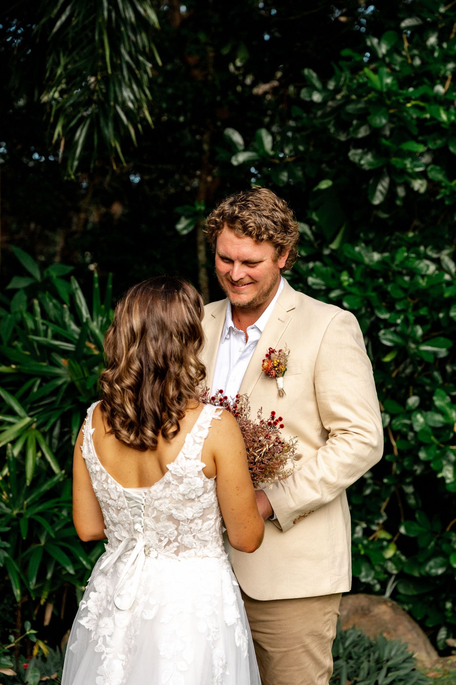 A smiling groom in a beige blazer gazes at his bride holding a dried floral bouquet outdoors.